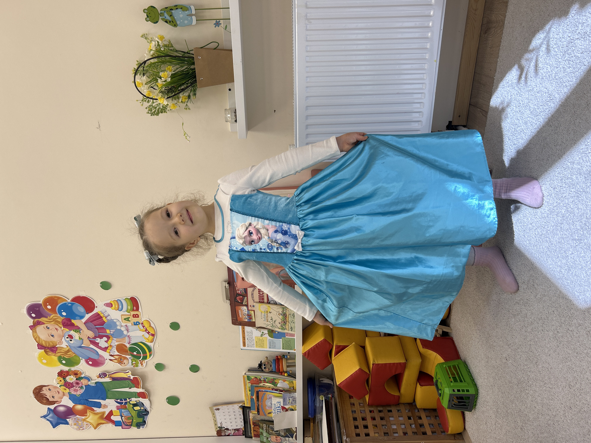 A young girl, approximately 4-6 years old, is captured indoors in what appears to be a child's room or play area in Izhevsk, Russia. She stands smiling directly at the camera, dressed in a light blue costume gown featuring an image of Elsa from Frozen on the bodice, worn over a white long-sleeved shirt and light pink socks. She playfully holds out the skirt of her dress.
The room is well-lit, suggesting daytime, with natural light casting shadows on the light-colored, carpeted floor. The cream-colored walls are decorated with cheerful, colorful wall stickers depicting a girl with balloons and a boy holding flowers, interspersed with smaller green circular decals.
To the viewer's left of the girl, shelves are filled with children's books, some with visible text including "МИРУ НАУЧИТЬСЯ," "АФРИКЕ," "Добрая," "mka," "Забавние ИСТОРИИ," and "РУГОСВЕТНОЕ ЕШЕСТР." A calendar with dates like "17 18 19 20" and "26 27" is also visible. Below the shelves, yellow and red soft play blocks are stacked alongside a green toy truck.
Behind the girl, a white radiator is affixed to the wall, and above it, a white shelf holds a brown paper bag containing artificial white and yellow flowers. Two decorative green frog figurines are placed above these items. The overall scene conveys a happy and playful atmosphere.