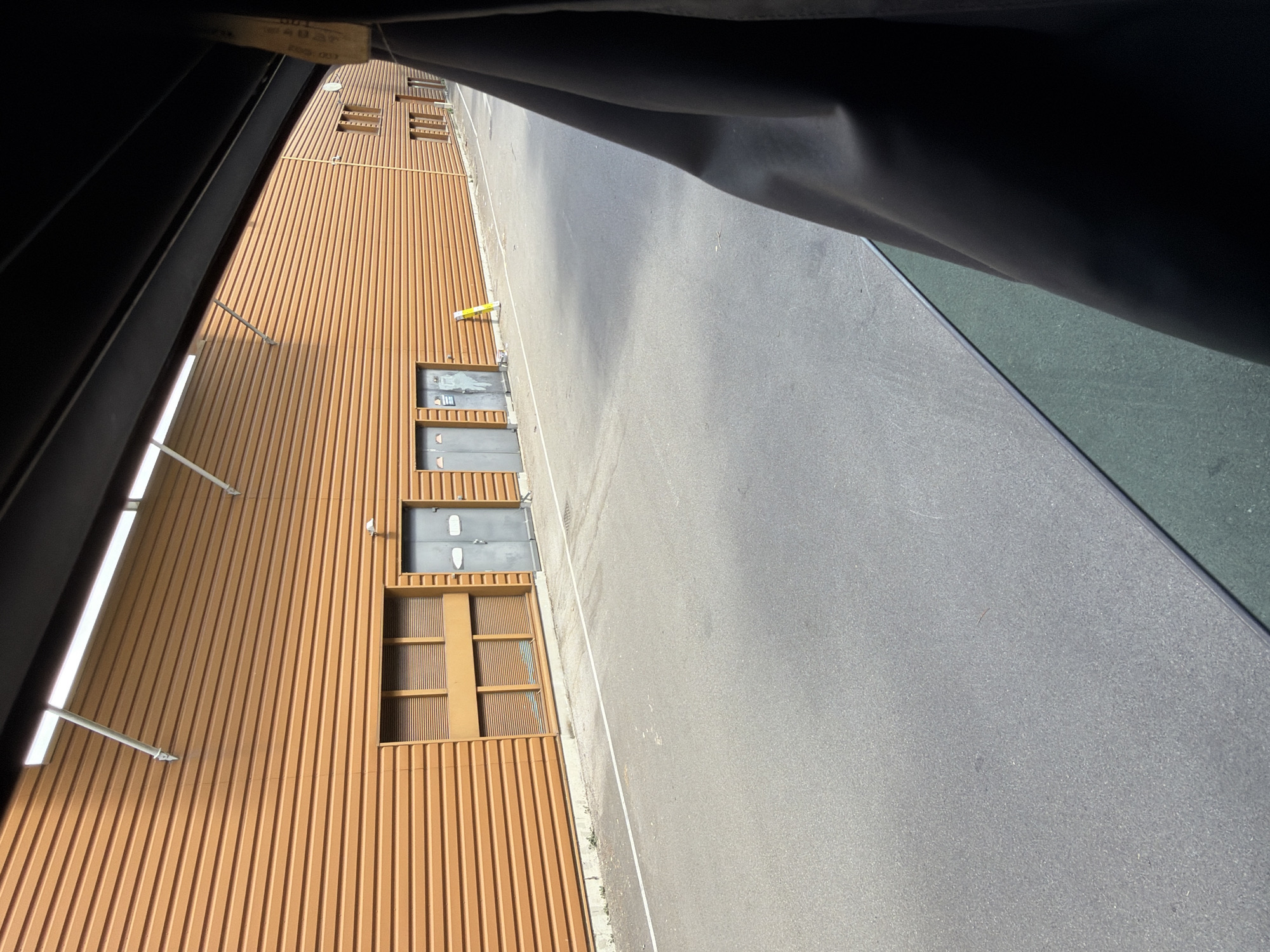 The image shows an outdoor scene in Arques, France, featuring a modern building with an orange corrugated metal facade. The building has several doorways, some with gray metal doors. To the left, there is a large window with louvers. The ground in front of the building is paved with asphalt and marked with a white line. A yellow bollard stands near one of the doors. The perspective suggests the photo was taken from inside a vehicle, with a dark fabric curtain obscuring the right side of the frame. The lighting indicates it is daytime.