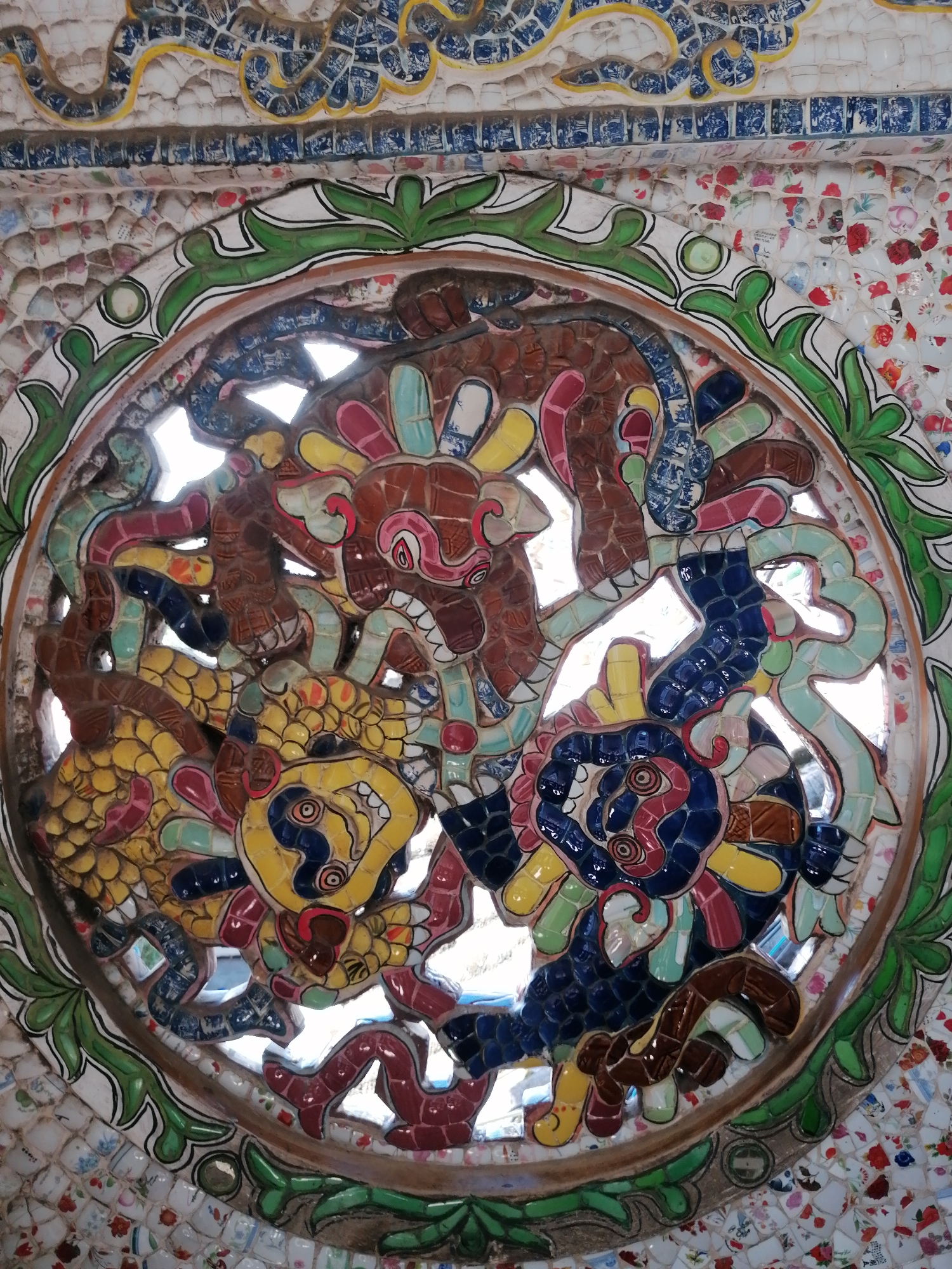 The image displays a circular mosaic artwork featuring mythical creatures, likely dragons, in vibrant colors. The mosaic is constructed from broken ceramic pieces. The background of the mosaic is white, with intricate patterns of blue and yellow tiling above and around the central circular artwork. The central artwork itself depicts several intertwined dragon-like figures in brown, yellow, blue, red, and green ceramic fragments. The dragons have distinct facial features with open mouths and sharp teeth. The surrounding area of the mosaic is composed of smaller, white ceramic pieces with colorful floral patterns, predominantly red and pink. The image is taken from a medium-close perspective, focusing on the detailed artistry of the mosaic. The location is identified as Lộc Quý, Vietnam.