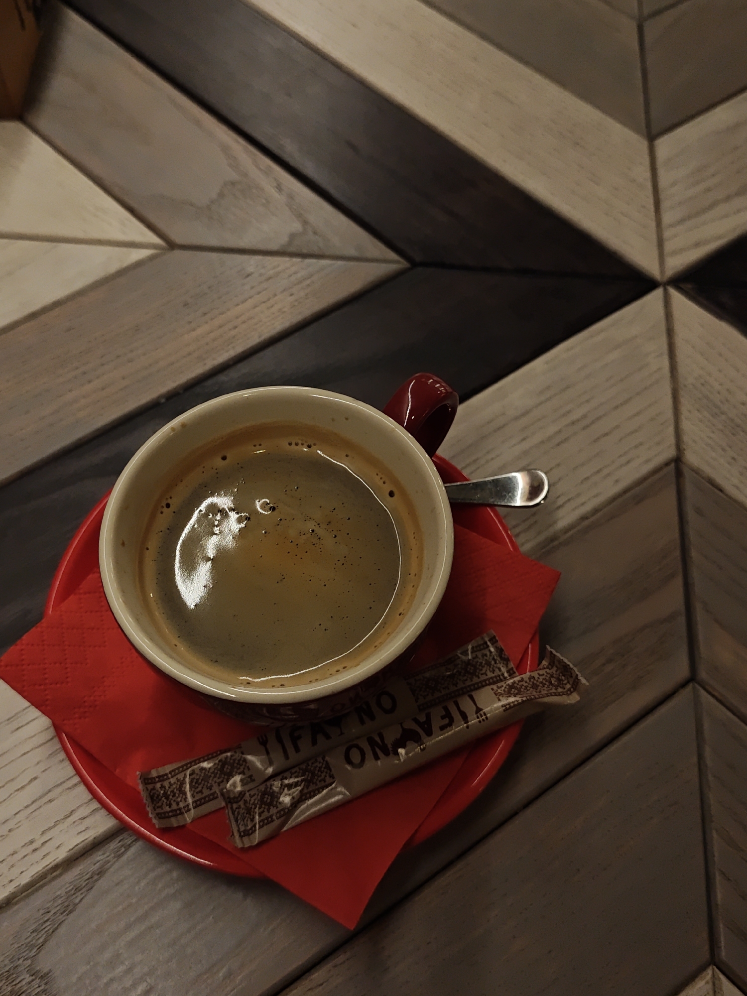A top-down shot depicts a cup of coffee on a red saucer, placed on a chevron-patterned wooden table. Next to the cup, on the saucer, are two sugar packets. A small spoon rests on the saucer. The scene is likely indoors, given the controlled lighting. The text on the sugar packets includes "IF AYNO" and "ON," but the rest is obscured. No people are visible in the image. Based on the text on the sugar packets, the location is likely in Ivano-Frankivsk, Ukraine.