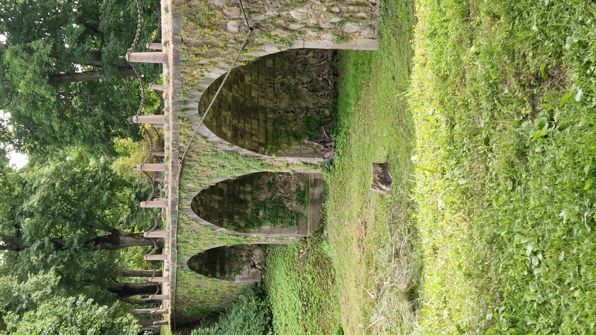 The image depicts an old stone bridge with three arched openings, covered in green ivy. The bridge is made of rough-hewn stone blocks and has stone posts with chains along its top. It is situated in a lush green park-like environment with tall trees in the background and dense vegetation and grass in the foreground. A tree stump is visible in the middle ground. The scene appears to be in Kazdanga, Latvia, likely within the grounds of the Kazdanga Manor. There are no people or animals present in the image, and no actions are being performed. The environment is natural and appears serene.