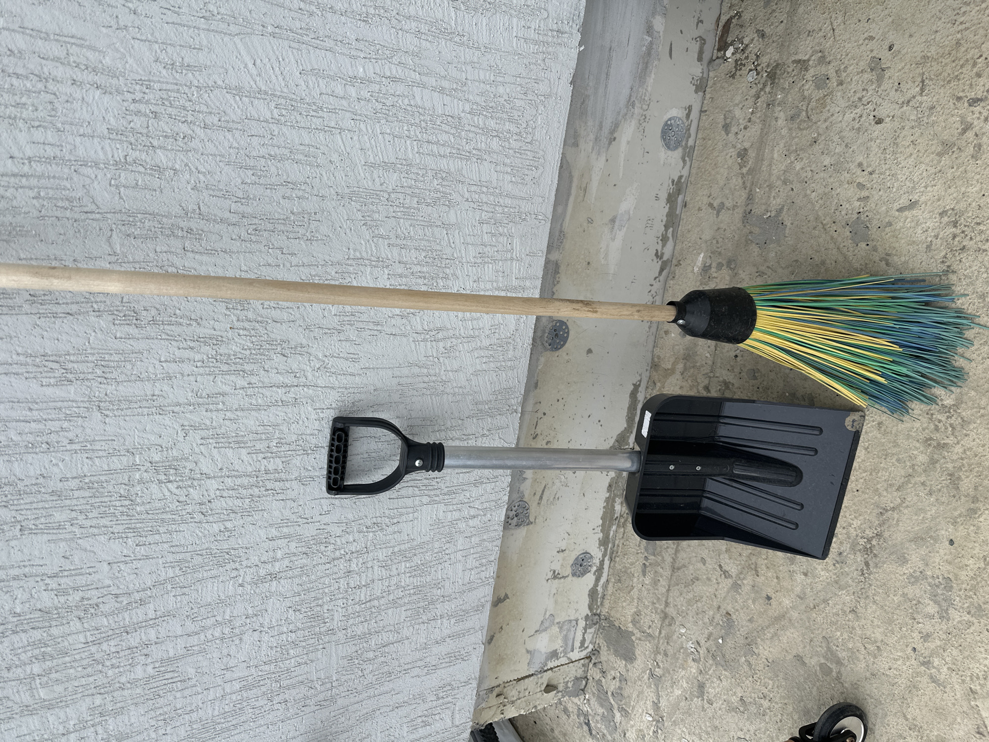 A broom and a shovel are laying horizontally on a concrete surface with a textured wall in the background. The broom has a wooden handle and bristles that are yellow, green, and blue. The shovel is black with a silver handle and a D-shaped black grip. The environment appears to be outdoors, possibly a patio or side of a building, in Unknown, Unknown. There are no people visible in the image.
