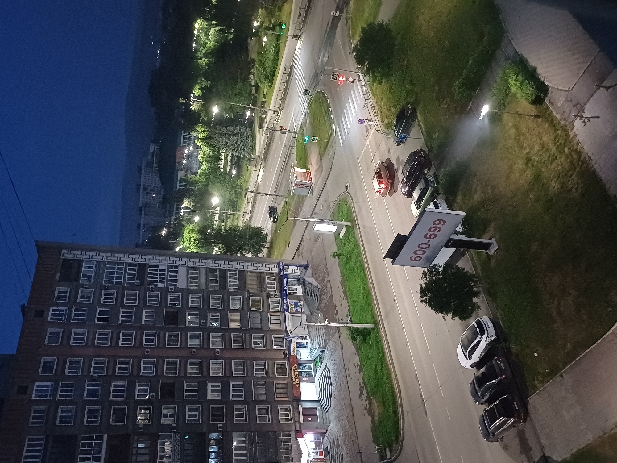 An elevated, medium shot captures a street scene at dusk or night in an urban environment. The primary subject is a tall, multi-story apartment building with numerous windows, its facade a dark, textured material. Below the building, a sidewalk runs alongside a street with visible lane markings. Several parked cars are present: a white sedan with damage to its rear bumper, two dark sedans, and a light-colored sedan further down the street. A billboard displaying "600-699" is positioned on the right side of the image. Across the street, a park area with trees and streetlights is illuminated, and a traffic intersection with pedestrian crossings and traffic signals is visible. In the background, distant hills and more buildings are silhouetted against the dark blue sky. The overall impression is a quiet, nocturnal urban landscape.