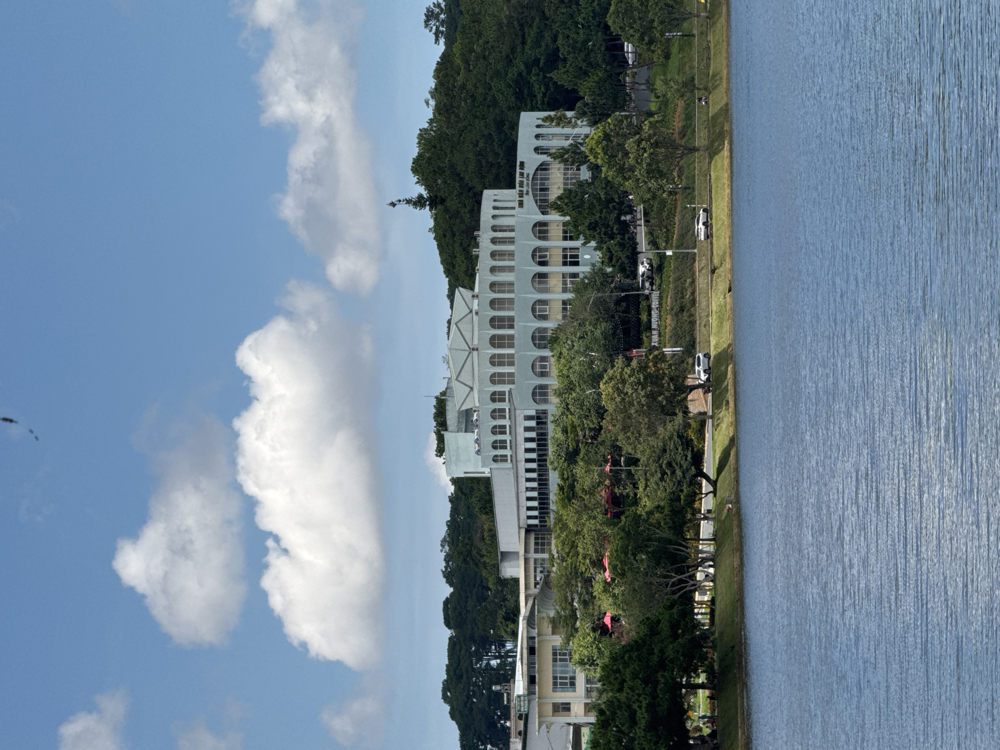 A wide shot captures Xuan Huong Lake in Da Lat, Vietnam, under a partly cloudy sky. The lake, a vibrant blue with gentle ripples, dominates the foreground. Beyond the lake, a lush green hillside is densely covered with trees. Situated on the hillside is a large, pale blue building with a distinctive arched facade and numerous windows. To the left of this building, a smaller, lighter-colored building with a flat roof is visible. A road runs along the edge of the lake, with several cars and a motorcycle present. Along the road, people are seen sitting on benches and walking in a park-like area with grass and trees. Some red umbrellas are visible amongst the trees. The overall scene depicts a serene, picturesque view of the lake and the surrounding cityscape.