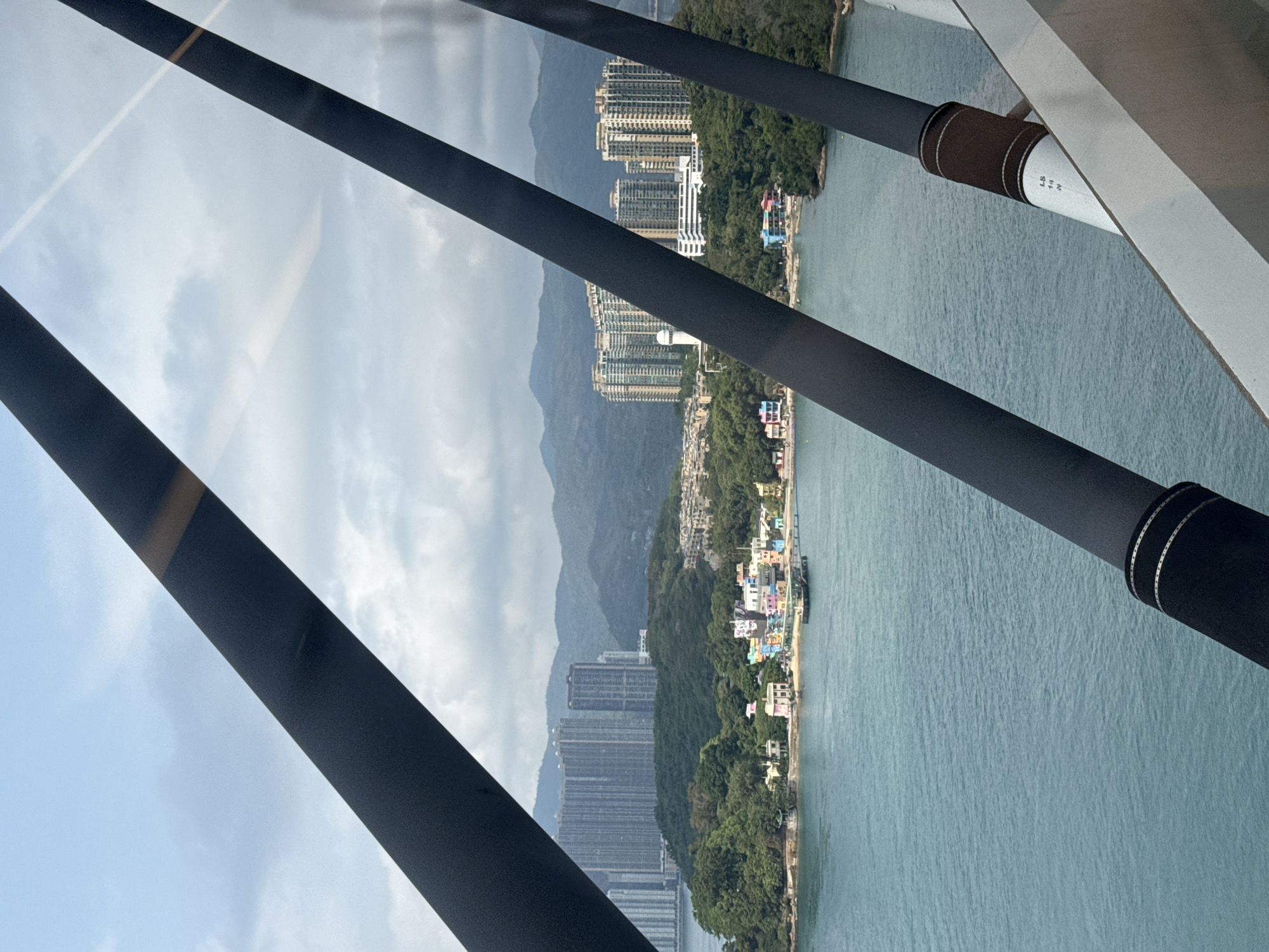 View from a cable car in Hong Kong, China, overlooking the shoreline with high-rise buildings, a mountain range, and clear water. The scene includes structural black beams, part of the cable car's construction, bisecting the landscape view. The environment appears bright with scattered clouds in the sky. The buildings vary in height and design, from residential towers to smaller structures along the water's edge. The shoreline is lined with greenery.