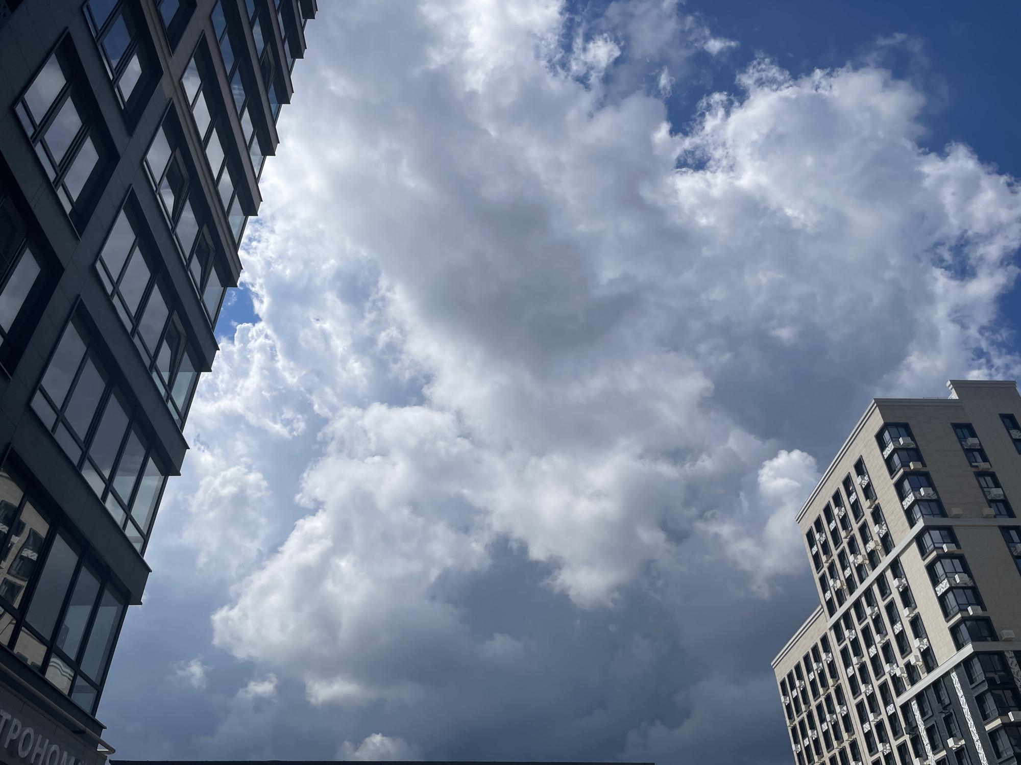 The image captures a low-angle view of modern apartment buildings against a dramatic sky filled with large, fluffy white and grey clouds. The building on the left is dark grey with numerous windows, angled towards the viewer. On the right, a lighter colored building with a patterned facade also features many windows and balconies. The scene appears to be in a city, likely Stavropol, Russia, given the architectural style and the cloudy sky often seen in the region. No people are visible in the image, and no actions or interactions are depicted. The focus is on the juxtaposition of the urban architecture with the dynamic atmospheric conditions.