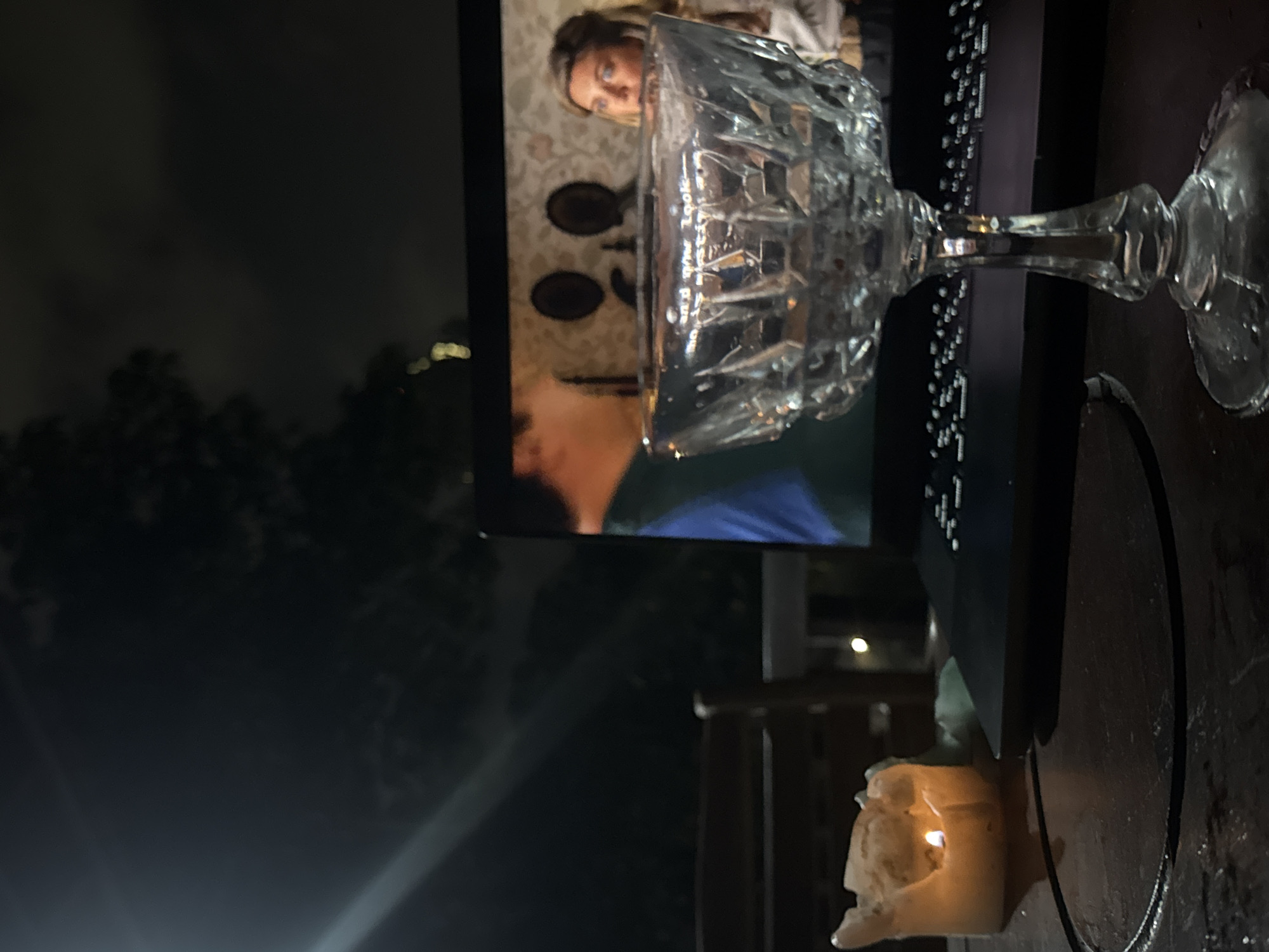 The image shows a dark outdoor scene at night, likely in a residential or semi-outdoor setting. The focus is on a laptop displaying a video of a woman indoors, with a crystal glass placed on the laptop's keyboard. A partially burned candle sits on a surface near the laptop, providing a dim light source. The laptop appears to be on a table or other flat surface. The background is obscured by darkness, with hints of foliage and possibly distant buildings. The image shows elements of a relaxed, informal setting. The specific location in Ungasan, Indonesia, is not discernible from the provided image.