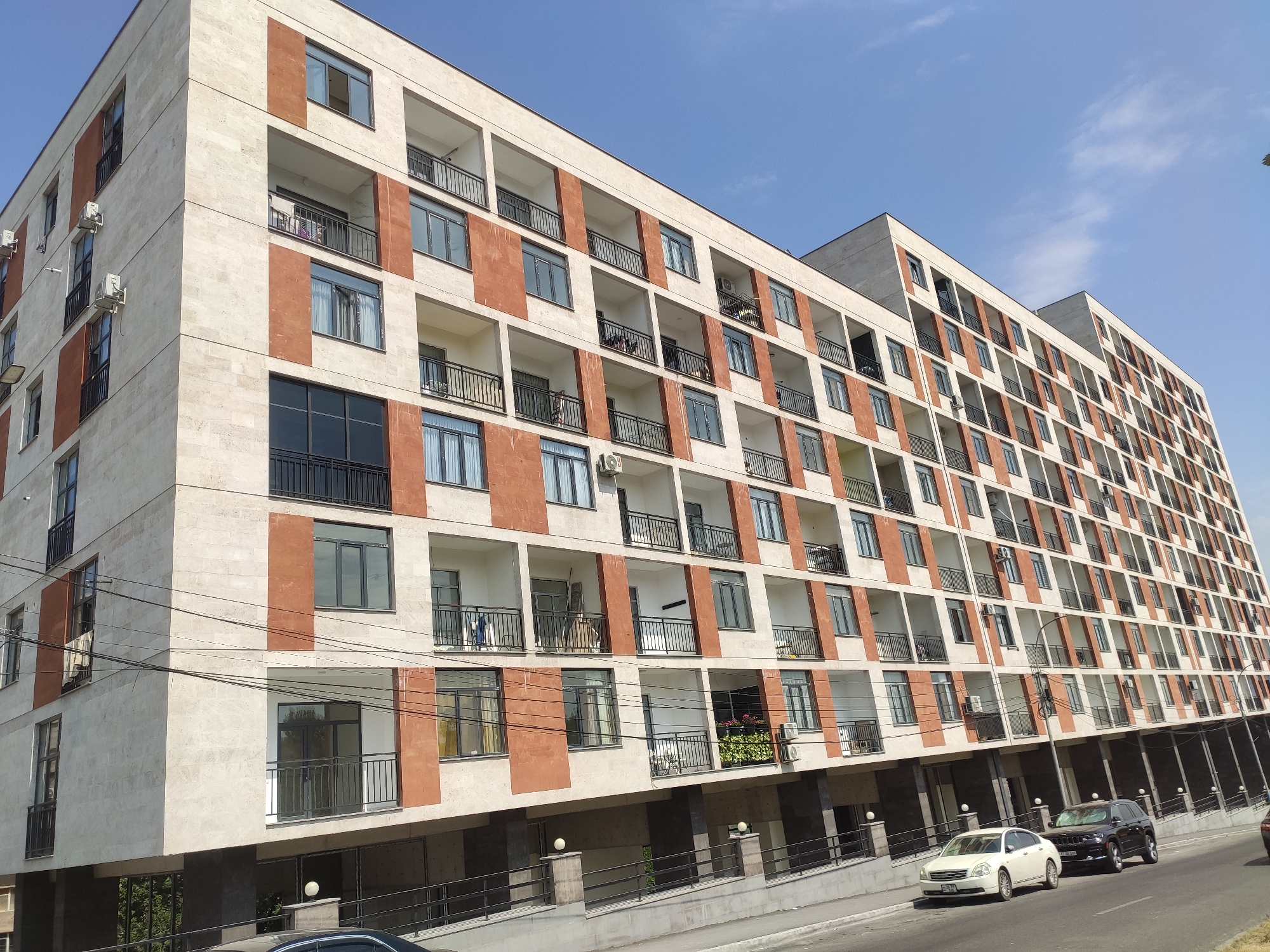 This is a medium-angle, eye-level exterior shot of a modern, multi-story apartment building with a light-colored stone facade and orange accent panels, located in Yerevan, Armenia. The building features numerous balconies with black railings and a repetitive pattern of windows. The sky above is clear with a few wispy clouds. In the foreground, a street runs alongside the building, with a light-colored sedan and a dark-colored SUV parked on the right side of the road. Electrical wires are strung across the facade of the building. The building's architecture suggests a recent construction or renovation, common in rapidly developing areas of Yerevan.