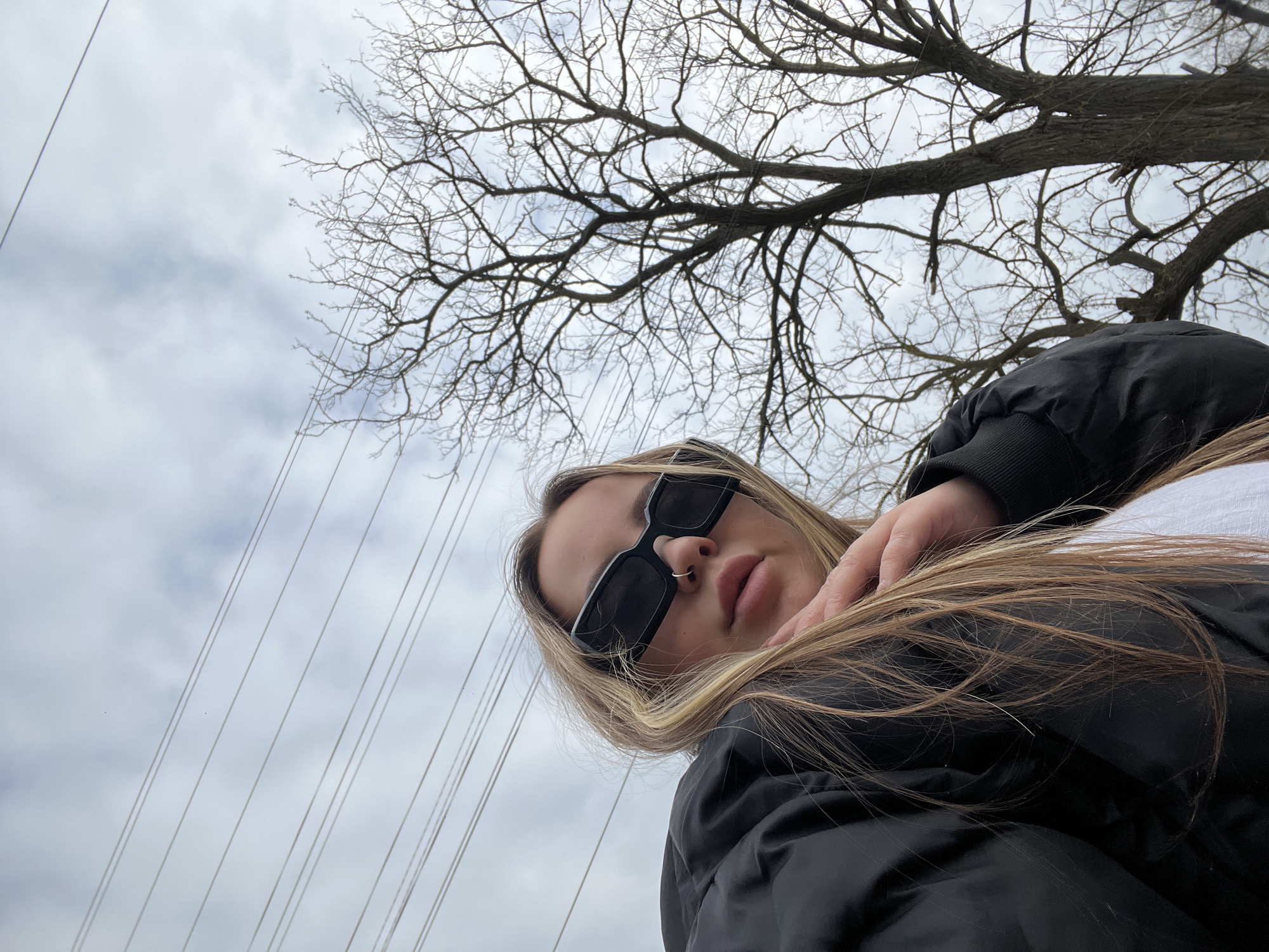 A low-angle outdoor shot features a young woman looking down towards the camera. She has long, straight blonde and light brown hair, which partially covers her black puffy jacket. Her face is partially obscured by dark, rectangular sunglasses, revealing a silver hoop nose ring and slightly parted lips. Her right hand is visible, resting near her left shoulder.
The background consists of an overcast sky with soft grey clouds. A large, leafless tree with a dense network of bare branches stretches across the upper and right side of the frame, suggesting an autumn or winter season. Several thin, light-colored wires or cables run diagonally across the sky from the upper left to the lower right. The overall light is diffused and soft due to the cloudy conditions. The woman's expression appears neutral and composed. The scene is set outdoors in Port-Katon, Russia, likely in an urban or semi-urban area given the presence of power lines. No visible text is present in the image.