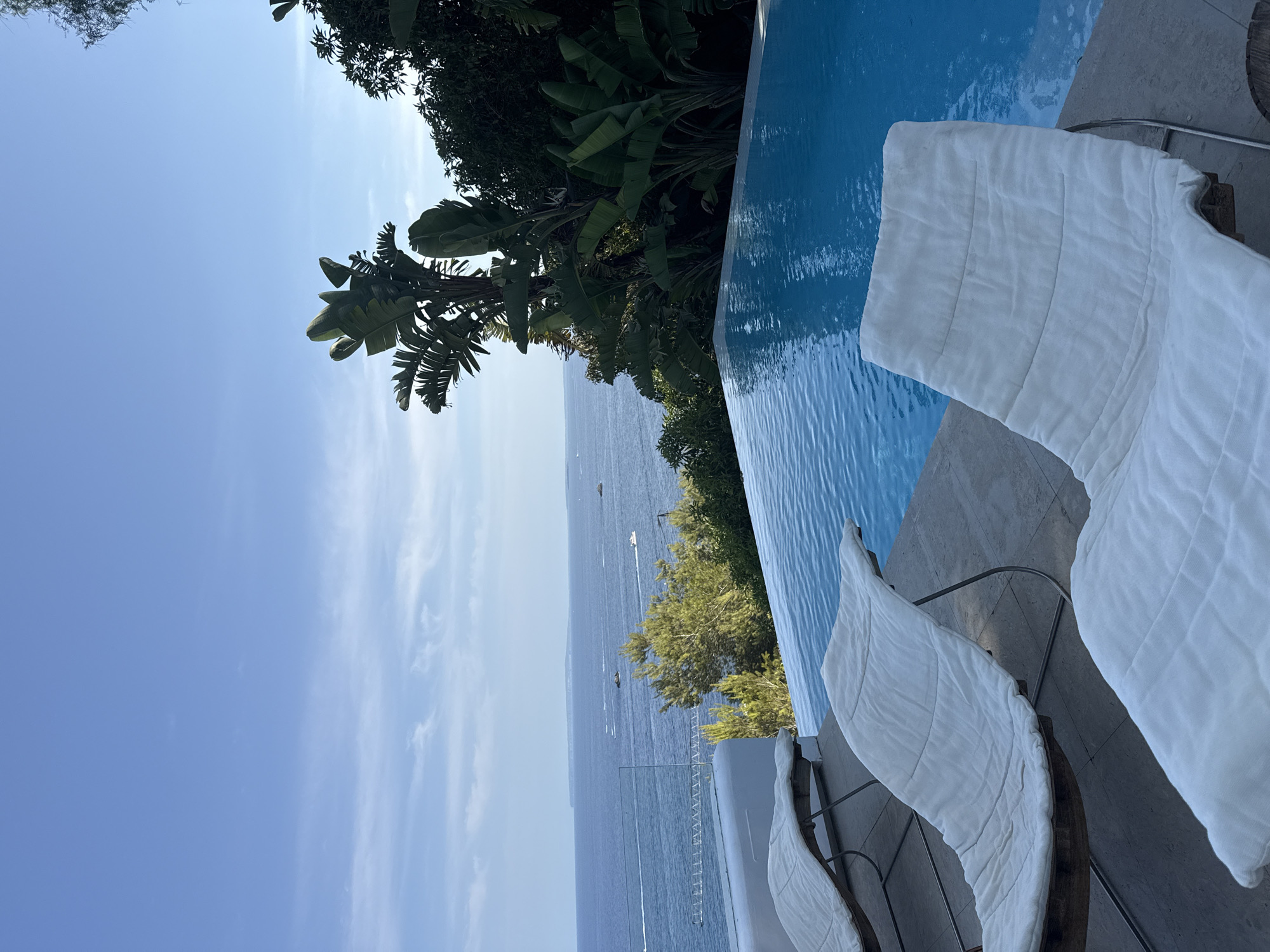 A tranquil infinity pool overlooks a calm blue ocean dotted with several small boats under a clear sky with scattered clouds. Lush green tropical foliage, including large-leafed plants, frames the right side of the scene. In the foreground, three white lounge chairs with plush cushions are arranged on a stone patio. The overall atmosphere is one of serene luxury.