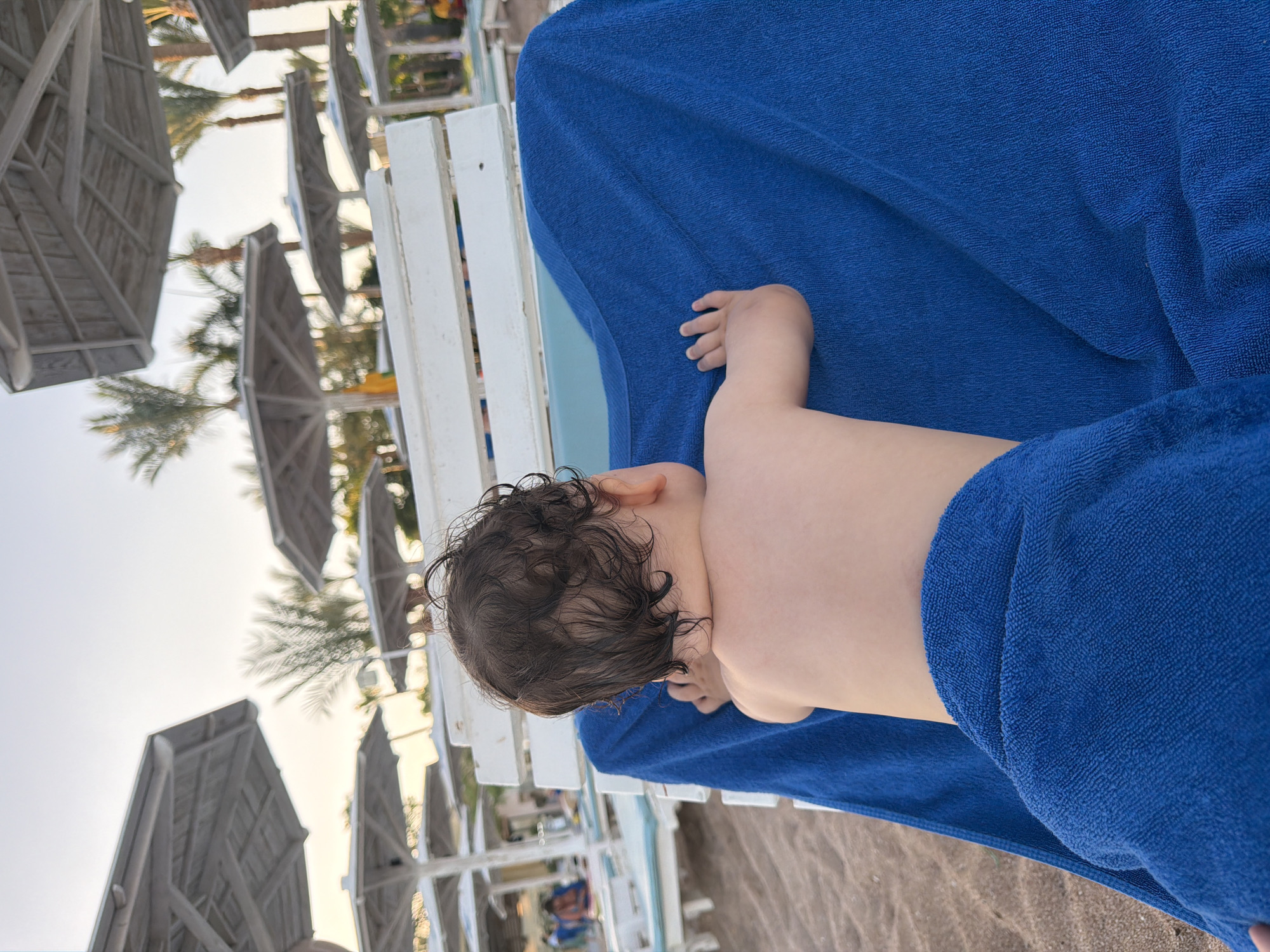 The media file shows a close-up of a baby lying on a sun lounger, covered by a blue towel, at a beach resort in Sharm El Sheikh, Egypt. The baby, seen from behind, has dark, wet hair and a small hand rests near its head. The background features rows of white sun loungers with their shades up, palm trees, and the overcast sky. The sandy beach is visible in the foreground. The overall impression is of a relaxed moment at a resort, possibly after a swim, given the baby's wet hair. The lighting suggests it is daytime, likely late afternoon or early morning due to the soft light. No visible text is present in the image.