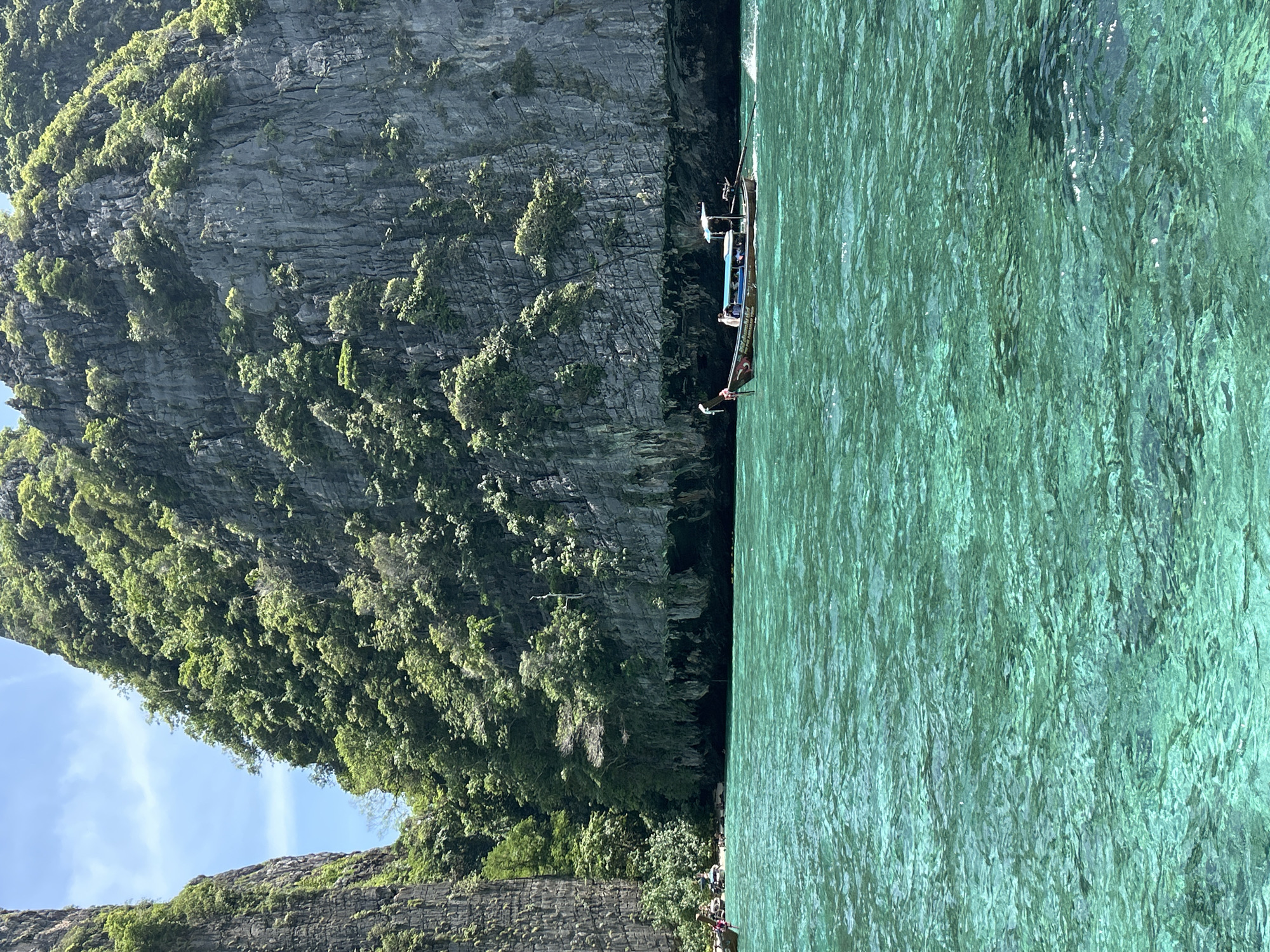 This image captures a serene scene in Ao Nang, Thailand, likely at Maya Bay or a similar location in the Phi Phi Islands, known for their towering limestone cliffs and emerald waters. The foreground is dominated by the vibrant turquoise sea, its surface rippled by gentle waves, revealing hints of the seabed beneath. A traditional longtail boat, painted with colorful decals, glides across the water, carrying several passengers and propelled by a man using a long oar. In the middle ground, a narrow stretch of beach is visible, dotted with a few more longtail boats anchored near the shore. A small group of people can be seen on the beach, enjoying the tropical environment. The background is characterized by immense, sheer limestone cliffs, densely covered in lush green vegetation. The cliffs rise dramatically from the water's edge, their textured surfaces indicating a long geological history. The sky above is a clear, bright blue with a few wispy clouds, suggesting a sunny day. The overall atmosphere is one of natural beauty and tranquility, typical of Thailand's stunning coastal landscapes.