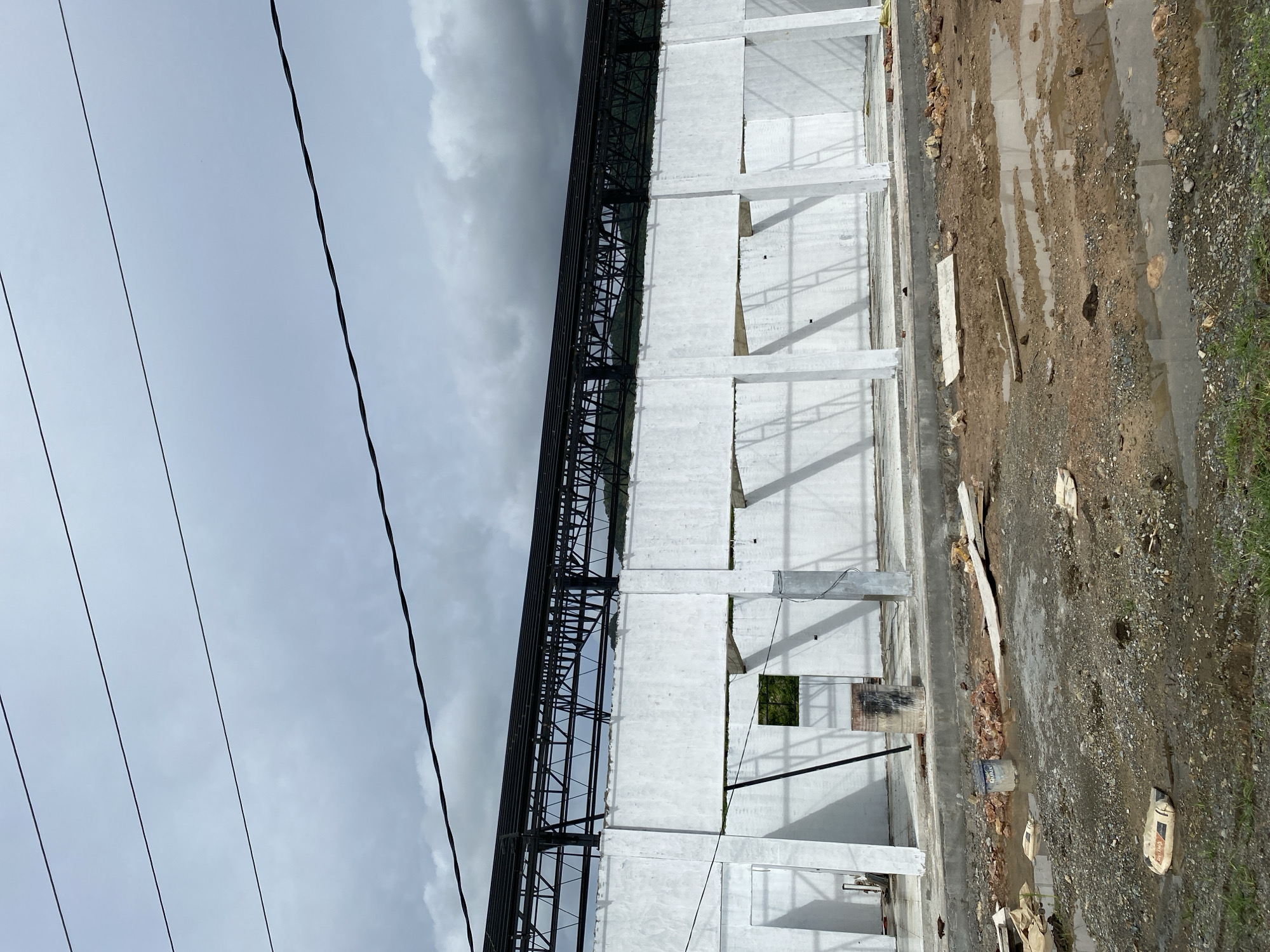 The image depicts an unfinished building structure with a white concrete facade and a black metal roof frame against a cloudy sky. The ground in front of the building is muddy with puddles and scattered construction debris, including bricks, wood planks, and bags of cement. There are four prominent power lines stretching horizontally across the upper portion of the image. The building appears to be a large, open structure with visible columns supporting the roof.