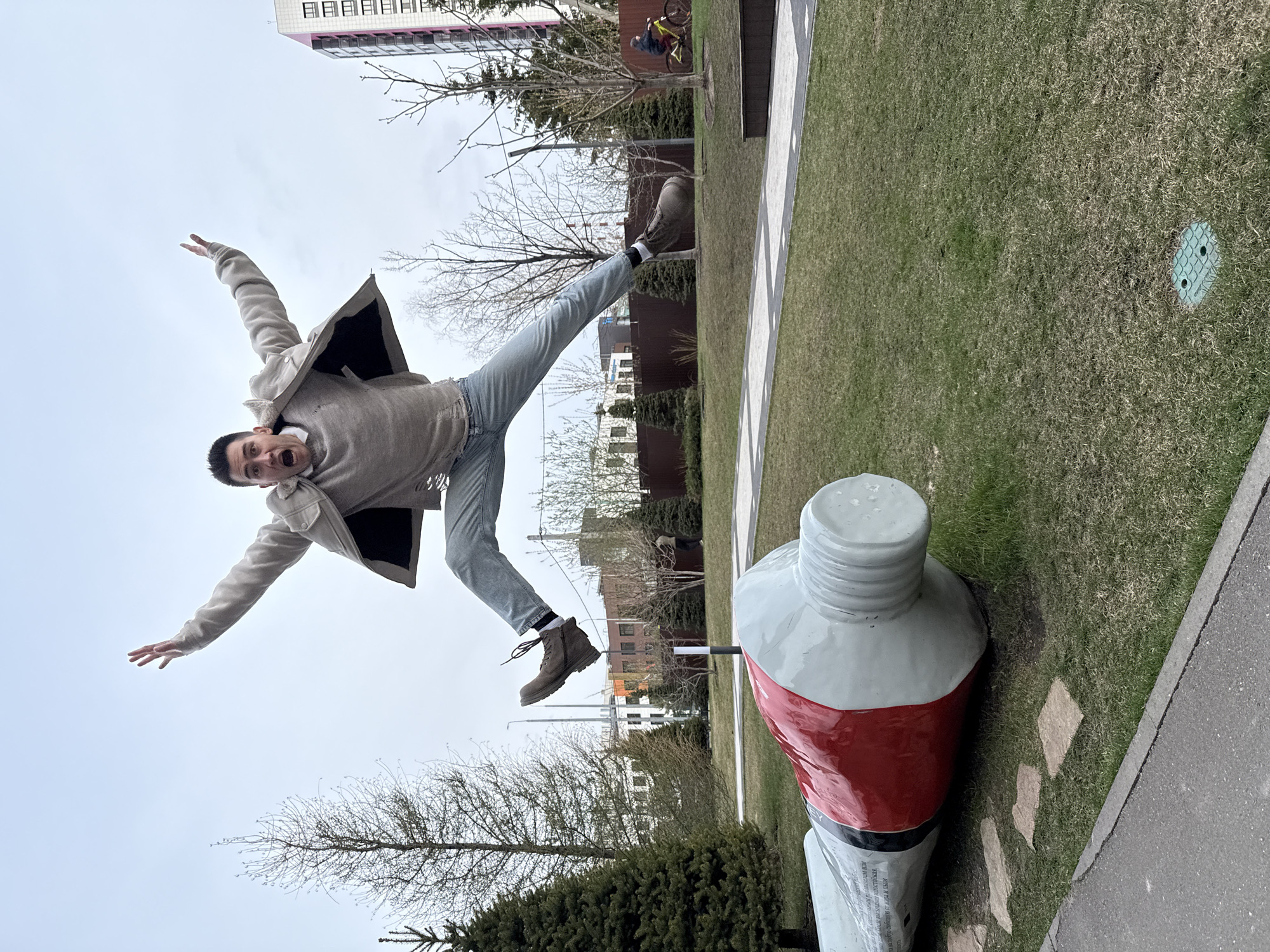 The media file, presented rotated 90 degrees clockwise, captures a dynamic outdoor scene in what appears to be an urban park in Kazan, Russia. A young man is prominently featured mid-air, having jumped, with his arms outstretched and legs splayed, conveying a sense of playful exuberance or surprise, evident from his wide-open mouth and eyes. He wears a light-colored jacket over a sweater and light blue jeans, paired with dark boots.
Below him, a large white and red sculpture resembling a giant tube of paint lies horizontally on a patch of green grass. A grey pathway runs alongside the grass, leading to stepping stones near the paint tube. In the background, there are bare deciduous trees, suggesting late winter or early spring, and a brown fence. Taller urban buildings, one with pink balconies, are visible against an overcast, bright sky. Further details include a green manhole cover in the grass and glimpses of other distant structures. Illegible text, possibly in Cyrillic, can be seen on the white body of the large paint tube sculpture.