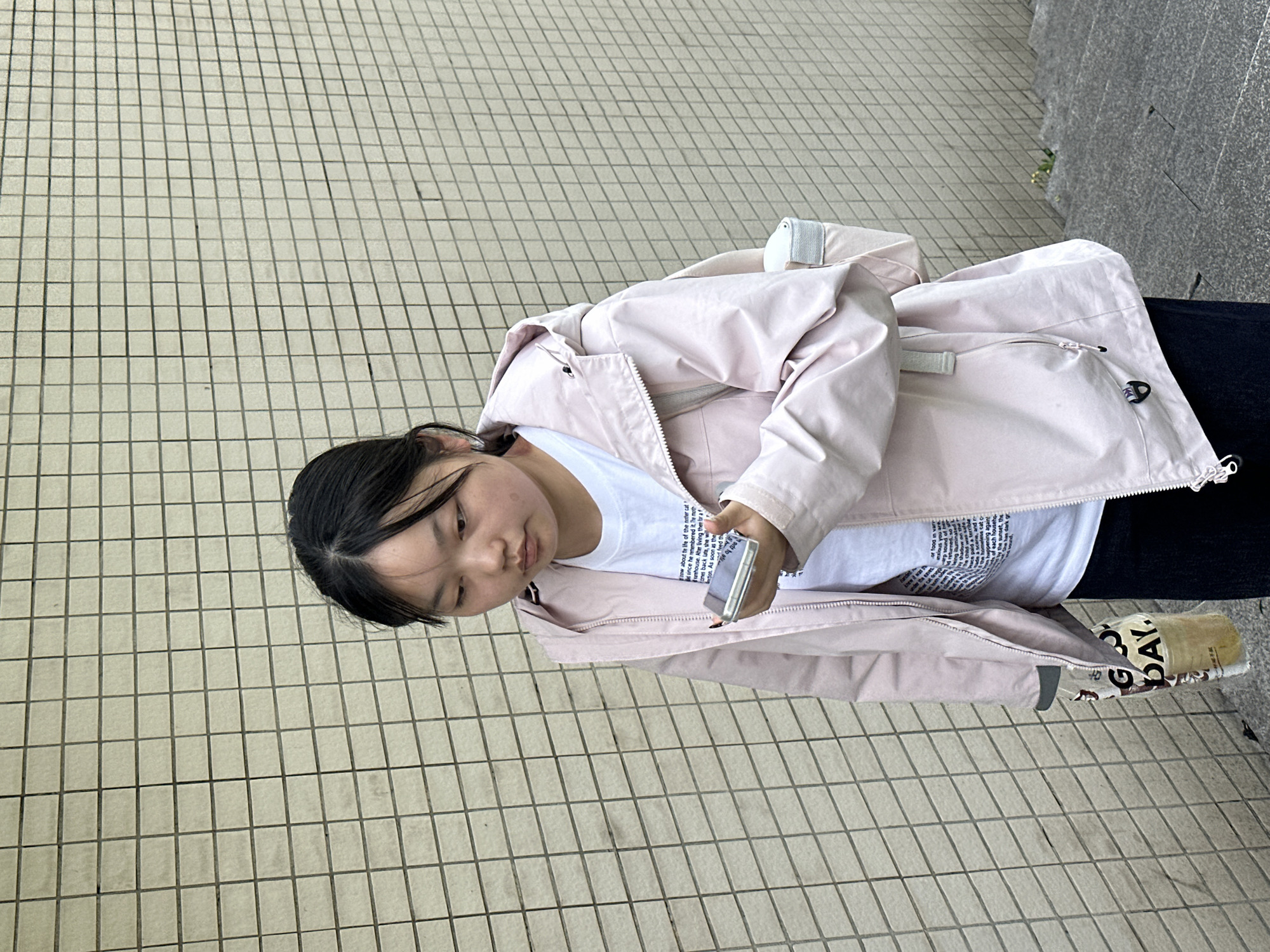 A young Asian woman is looking down at a smartphone she holds in her right hand, while standing on a paved surface. She wears a light pink jacket over a white t-shirt and dark pants. Her hair is black. A “GU DAY” bag is visible on the ground near her right foot. The location is potentially in the city of Jiangjin, China. The backdrop consists of a tiled floor or walkway and a textured gray wall.