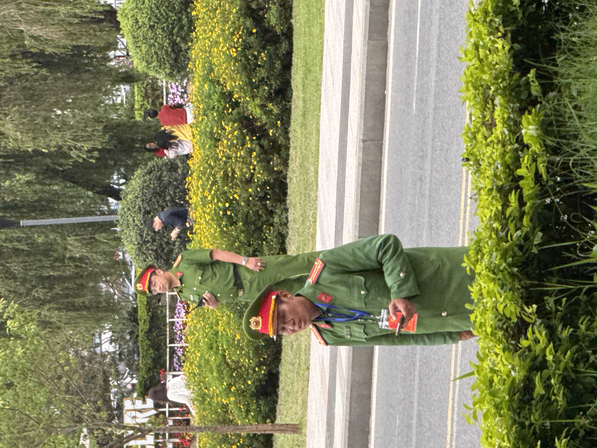 Two uniformed male officers are standing in the foreground, with a blurred background of people and greenery. The officer on the left is closer to the camera and is wearing a green uniform with a red-trimmed cap. He is holding a red identification card and a cell phone. The officer on the right is standing further back, also in a green uniform, and is holding a black object, possibly a radio. In the background, there are several people seated and standing near a bush and a flowerbed. The scene appears to be outdoors in an urban environment, with a road and sidewalk in the foreground. The presence of the officers suggests a public event or security detail. The foliage and the style of the uniforms are consistent with locations in Southeast Asia. Given the context, this scene could be situated near a public space in Da Lat, Vietnam, such as a park, plaza, or along a main street.