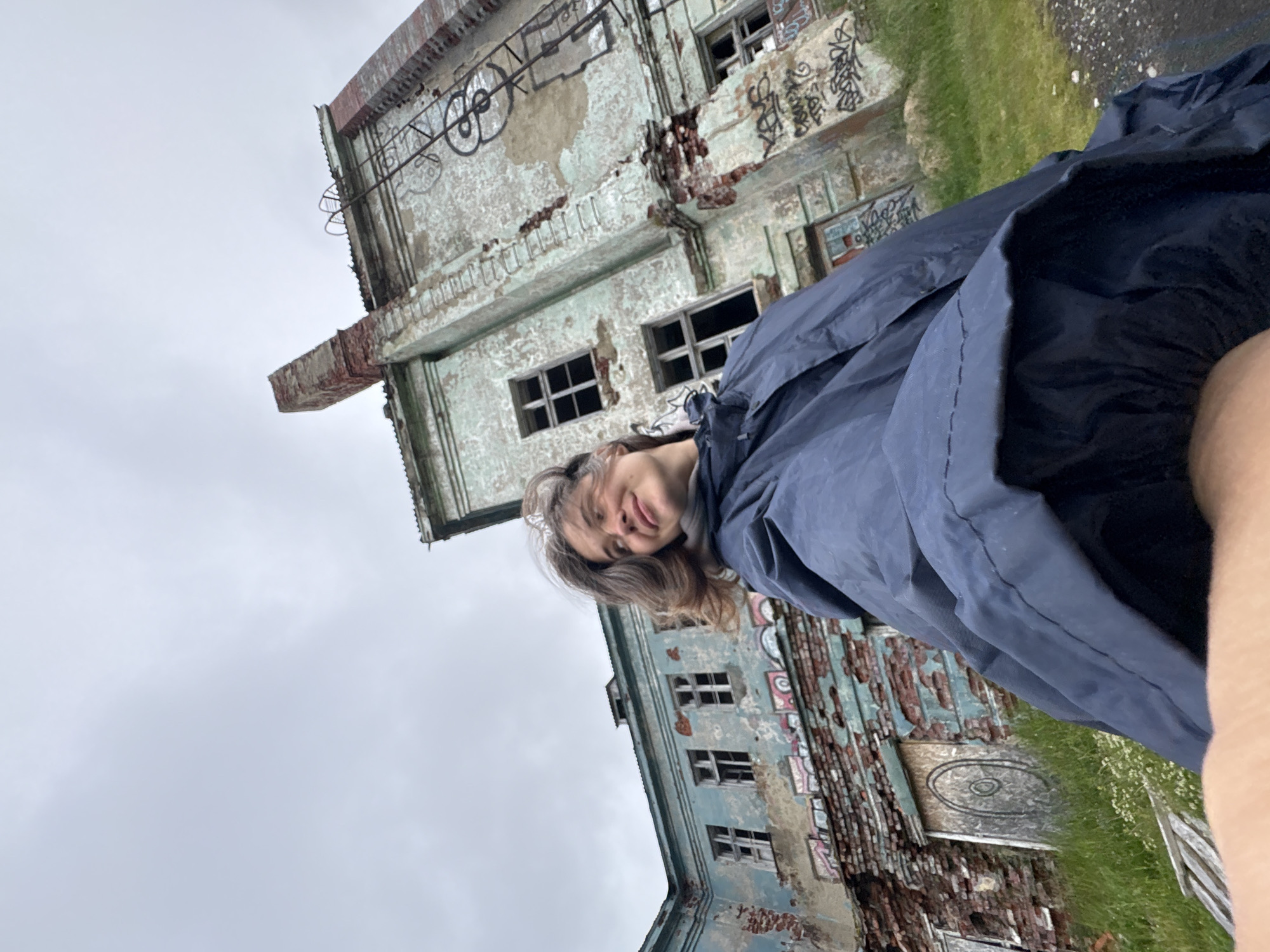 A low-angle shot captures a young woman wearing a dark blue rain jacket. She is looking upwards and to the right with a slight smile, her hair gently blowing. Behind her, an abandoned, weathered building with peeling blue paint and exposed brick stands against an overcast sky. The structure features several broken windows, graffiti, and a prominent brick chimney. The building appears to be located in Teriberka, Russia, a coastal village known for its abandoned industrial sites and harsh, northern climate. The ground in front of the building is covered in patchy green grass and some white wildflowers.