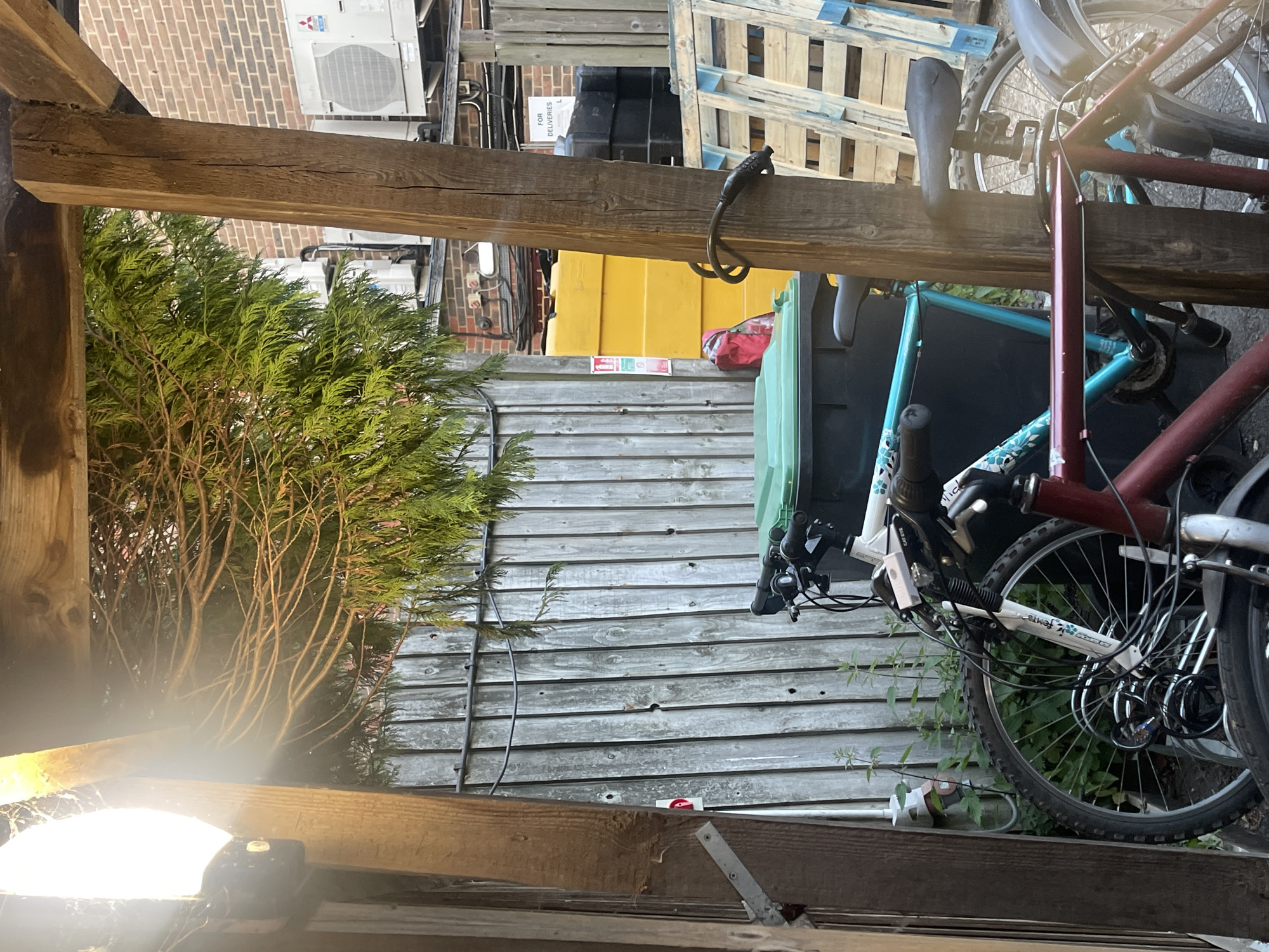 The image depicts an outdoor storage or cluttered area in Maidstone, UK, likely a backyard or side alley. A wooden structure frames the view, with a beam prominent in the foreground. A dense evergreen bush partially obscures a weathered wooden fence or siding. A stack of wooden pallets is visible, along with a yellow container and a black bin. Bicycles are stacked or leaned against the structure, secured with a lock. Electrical wiring is seen along the wooden siding. An air conditioning unit is mounted on a brick building in the background.