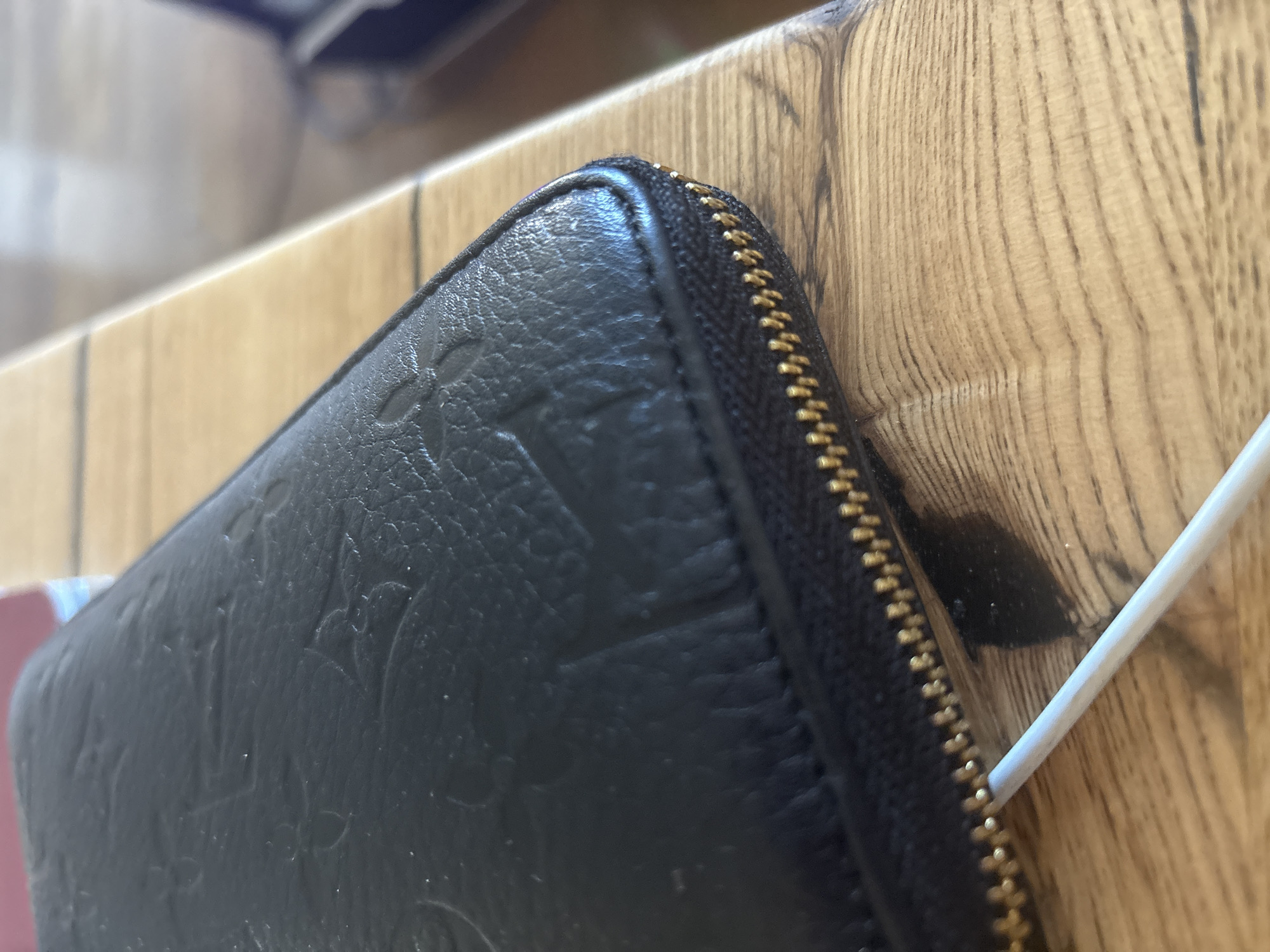 A black embossed wallet is resting on a wooden surface, with a white cable extending from beneath the zipper. The wallet exhibits a patterned texture with embossed floral or monogram motifs. The wooden surface shows visible grain and appears to be a table or countertop. The background is blurred, but suggests an indoor environment. The image was captured in the city of Orekhovka, Russia.