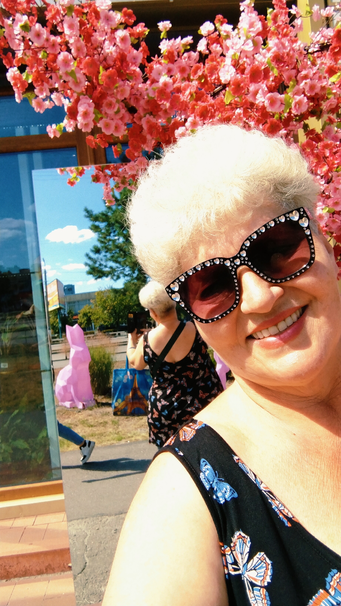 In a selfie taken in Horishni Plavni, Ukraine, a woman with short, light-colored hair and wearing bejeweled sunglasses smiles. She is dressed in a black dress with a butterfly print. In the background, reflected in a large mirror, another woman is visible, also wearing a butterfly-print dress and holding a camera. She appears to be taking a picture. A large, pink, geometric rabbit sculpture stands to the left of the reflected woman. Pink cherry blossoms are visible in the upper portion of the image, framing the scene. The environment appears to be outdoors, with a blue sky and some trees visible in the reflection.