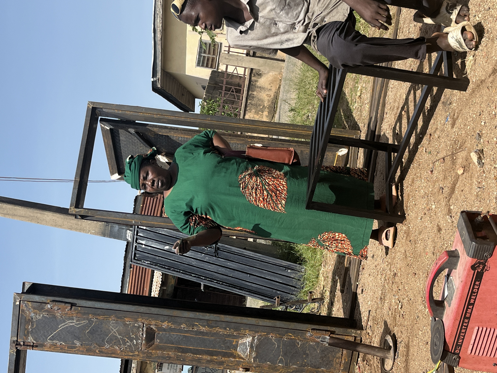 The image depicts an outdoor scene in the city of Jos, Nigeria, characterized by a dusty, sun-drenched environment. In the foreground, a red DC inverter MMA welder sits on the ground. A young person wearing a light-colored, short-sleeved shirt and dark pants is partially visible on the right, seated with their legs extended and resting on a black metal frame. They are wearing light-colored slides on their feet and yellow sunglasses pushed up onto their head.
In the center of the image, a woman stands dressed in a vibrant green dress with an orange and black patterned fabric at the hem and upper chest area. She wears a green headscarf and light-colored sandals. Her left arm is bent, and she is gesturing with her left hand, appearing to be in mid-conversation. A brown bag hangs from her right shoulder.
The background is populated with various objects, including large, rusty metal door frames leaning against each other. One door frame is particularly prominent on the left. Further back, there are corrugated metal roofs of buildings, suggesting a semi-urban or workshop setting. The ground is covered with loose gravel and dirt, with some patches of dry grass visible. The sky is clear and blue, indicating fair weather. The overall impression is of an active workspace, possibly a metal fabrication or repair shop, under a bright sky.