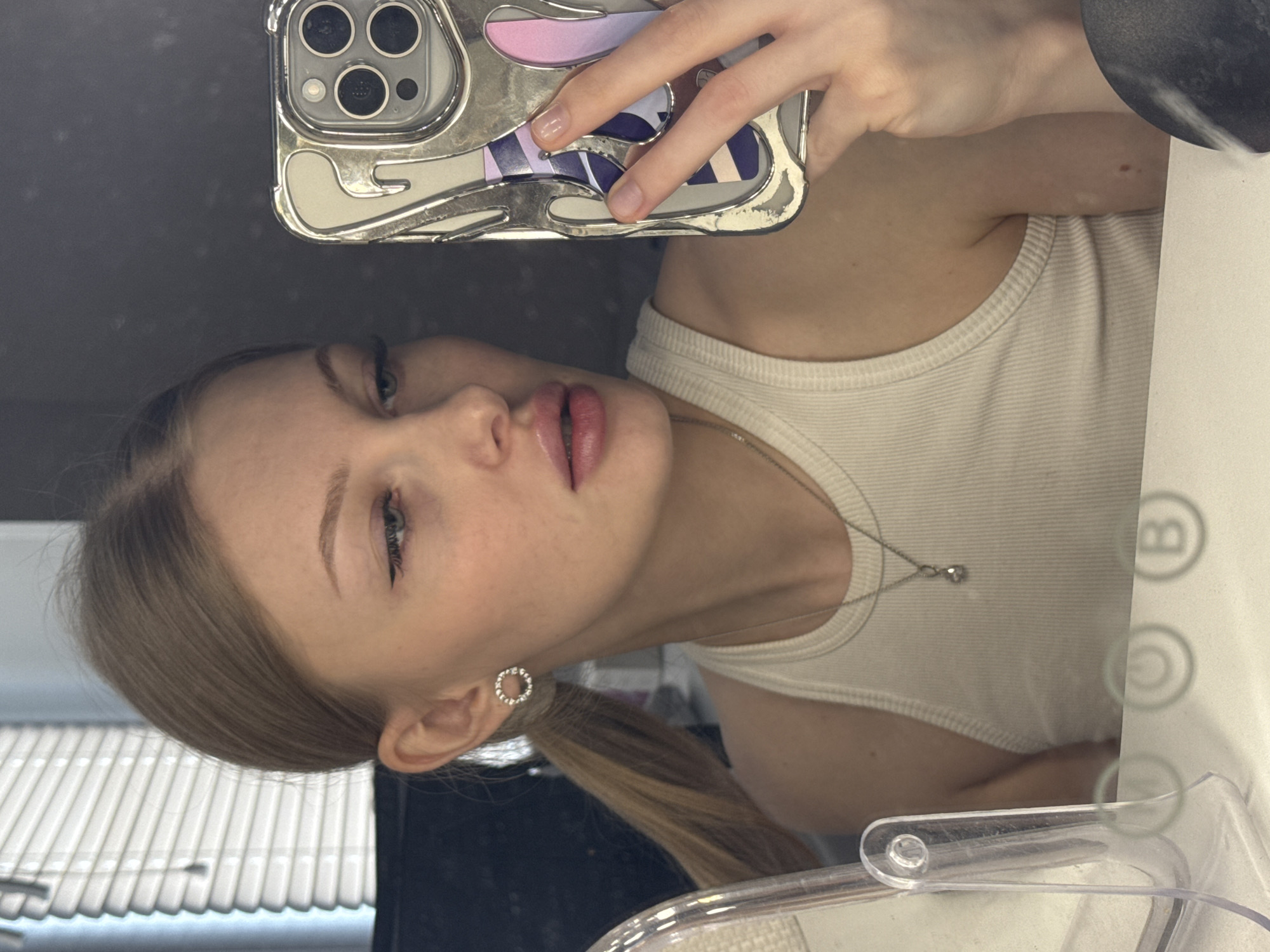 A young woman takes a selfie in what appears to be a bathroom. She is holding an iPhone in her right hand with a silver-colored case that has a decorative pattern. Her phone obscures the upper part of her head. She wears a white tank top and a delicate silver necklace with a small pendant. Her blonde hair is pulled back into a low ponytail, and she has circular silver earrings. The background includes a window with white blinds and a vanity area with a mirror and lighting controls labeled with "B", "C", and other illegible symbols. The location is impossible to determine based on visual evidence.