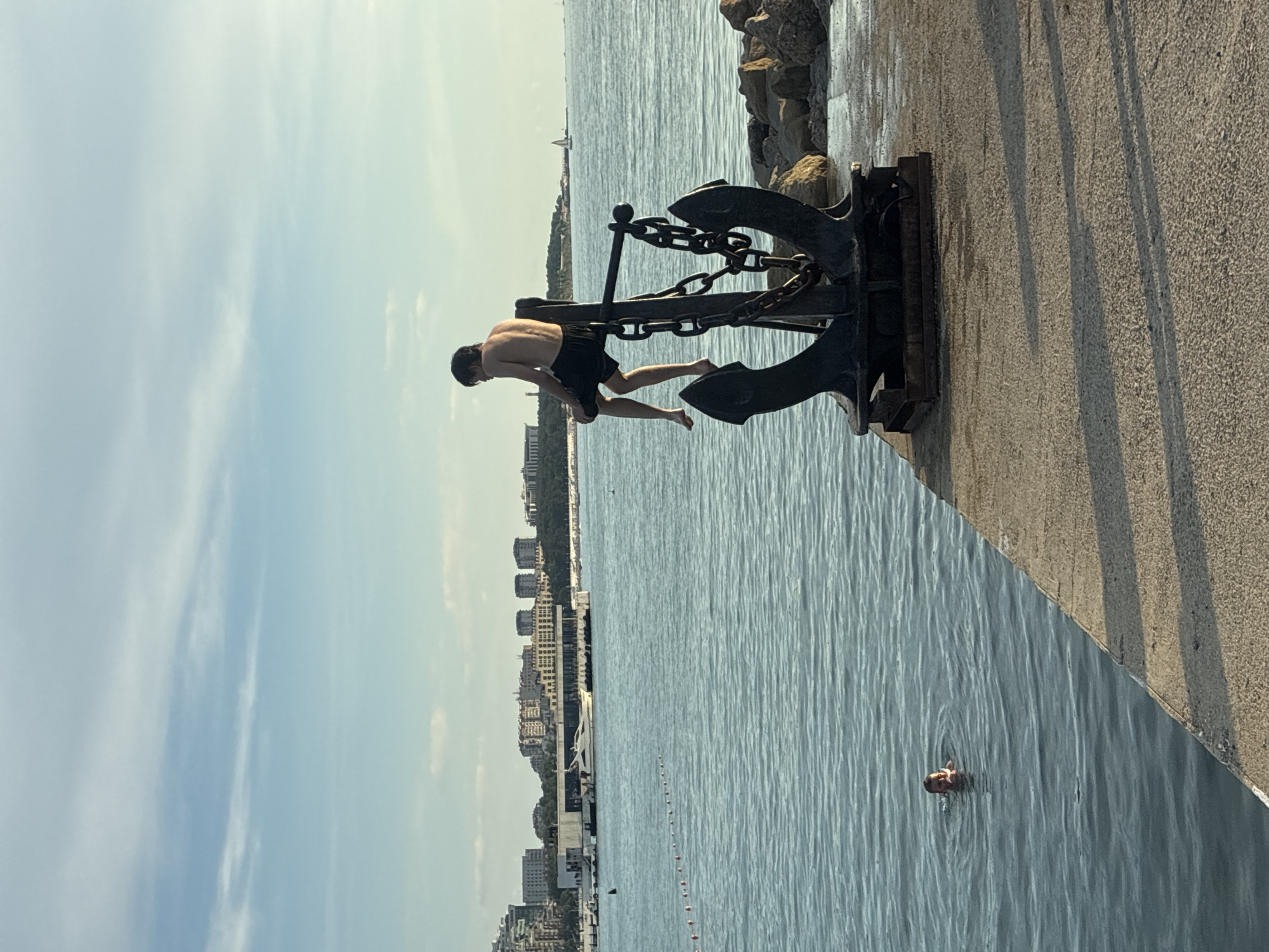 A young male, shirtless and wearing black shorts, sits on top of a large, black, rusted anchor with a thick chain attached. He is positioned on the edge of a concrete pier, with his legs dangling over the water. In the middle-left of the frame, another person's head is visible above the surface of the blue water, suggesting they are swimming. The background reveals a cityscape with multiple buildings and a clear blue sky with scattered clouds. The sea extends to the horizon, where a sailboat can be seen in the distance. The pier is made of concrete and has a rough texture, with shadows cast by the anchor and an unseen object. The scene appears to be a waterfront area in Gelendzhik, Russia, likely a harbor or promenade.