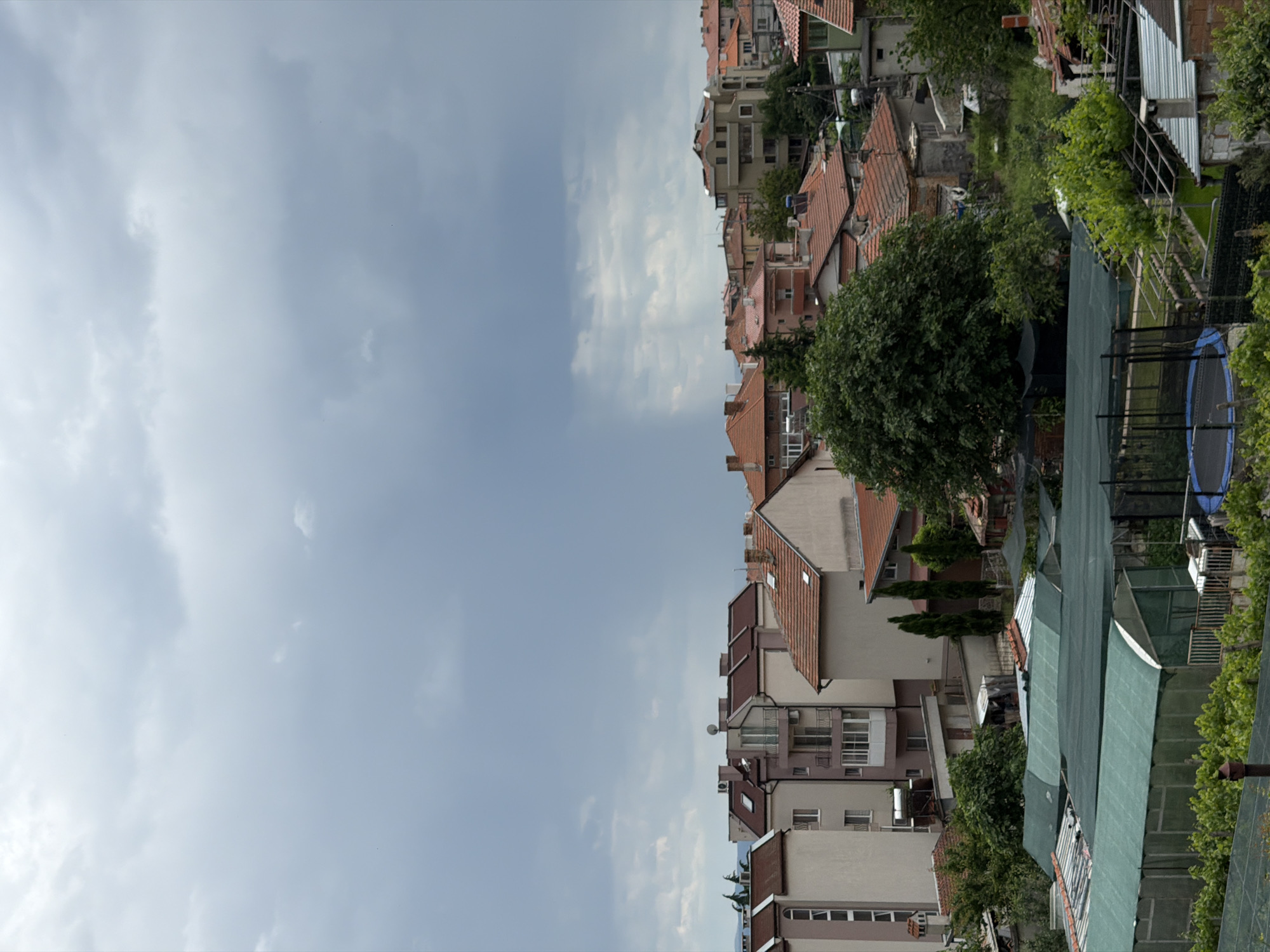 The image shows a cityscape scene, likely in Burgas, Bulgaria, judging by the architecture style of the buildings. The sky is overcast with diffused light, with clouds dominating the left side of the image. The right side is dominated by residential buildings of varying heights, mostly with terracotta-tiled roofs. There's significant greenery interspersing the buildings, suggesting residential gardens or trees along the streets. In the foreground, there is a trampoline and some metal fences.