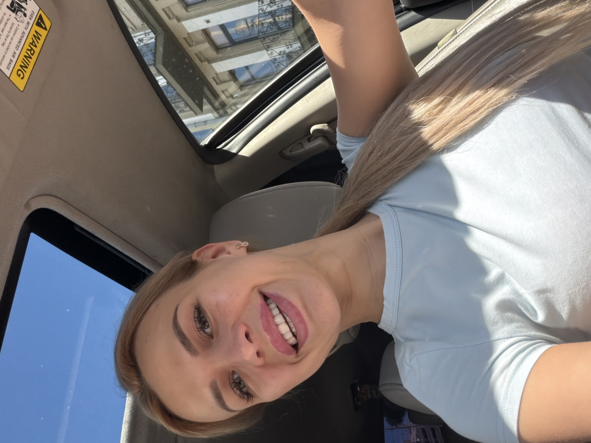 The image shows a young woman with blonde hair inside a car, with her head tilted back and looking up towards the sunroof. She is smiling broadly, with her tongue sticking out slightly. She is wearing a light blue t-shirt and has a small earring in her left ear. The sunroof of the car is open, revealing a blue sky and some buildings reflected in the glass. The interior of the car is visible, including the sun visor with a warning label. The sun visor has a yellow warning sticker with black text that reads "A WARNING", "EVEN WITH ADVANCED AIR BAGS", and "Children can be killed or seriously injured by the air bag". The sticker also mentions "The back seat is the safest place for children". The lighting suggests it is daytime, likely sunny. The visible buildings in the reflection hint at an urban environment. The overall mood of the image is playful and cheerful. The location context is Kyiv, Ukraine.