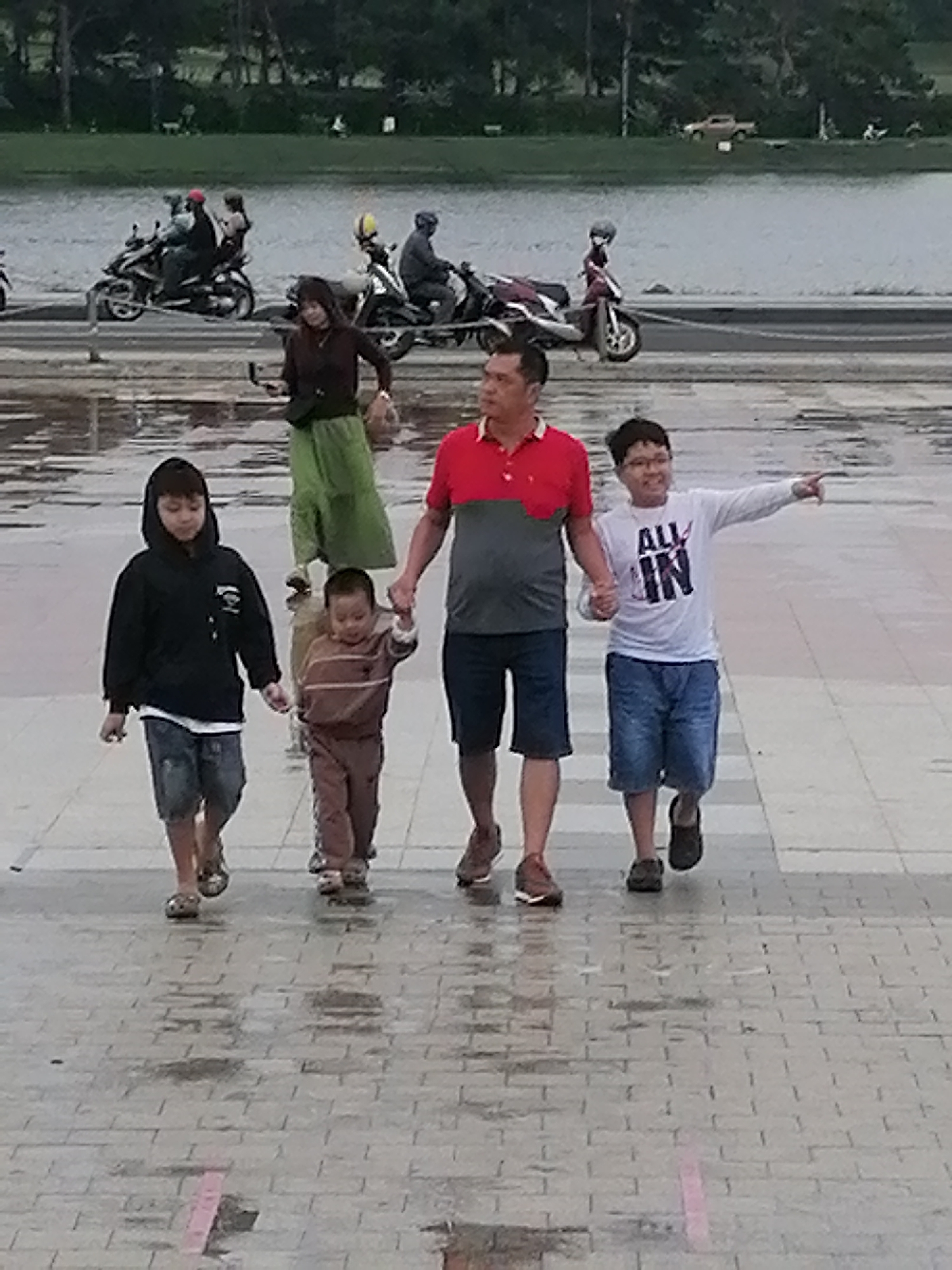 A group of five people, two adults and three children, are walking on a wet, paved walkway. The walkway is bordered by a body of water in the background, with trees and a hillside behind it. The group is walking from left to right in the frame.
The adult on the left is a man wearing a red and gray polo shirt and dark shorts. He is holding hands with a young boy on his left, who is wearing a brown striped long-sleeved shirt and pants.
To the left of the boy is another child, a young boy, wearing a black hooded sweatshirt and denim shorts. He is also holding hands with the boy in the brown outfit.
To the right of the man is a teenage boy wearing a white t-shirt with black lettering that says "ALL IN" and blue denim shorts. He is pointing to the right.
In the background, several motorbikes are parked along a road. One motorbike has a woman in a dark top and a long green skirt on it, holding a phone. Another motorbike has a person in a red helmet. The scene appears to be in a public area, possibly a park or a waterfront promenade in Da Lat, Vietnam, given the presence of motorbikes, the landscape, and the overall atmosphere. The ground is wet and reflects light, suggesting recent rain.