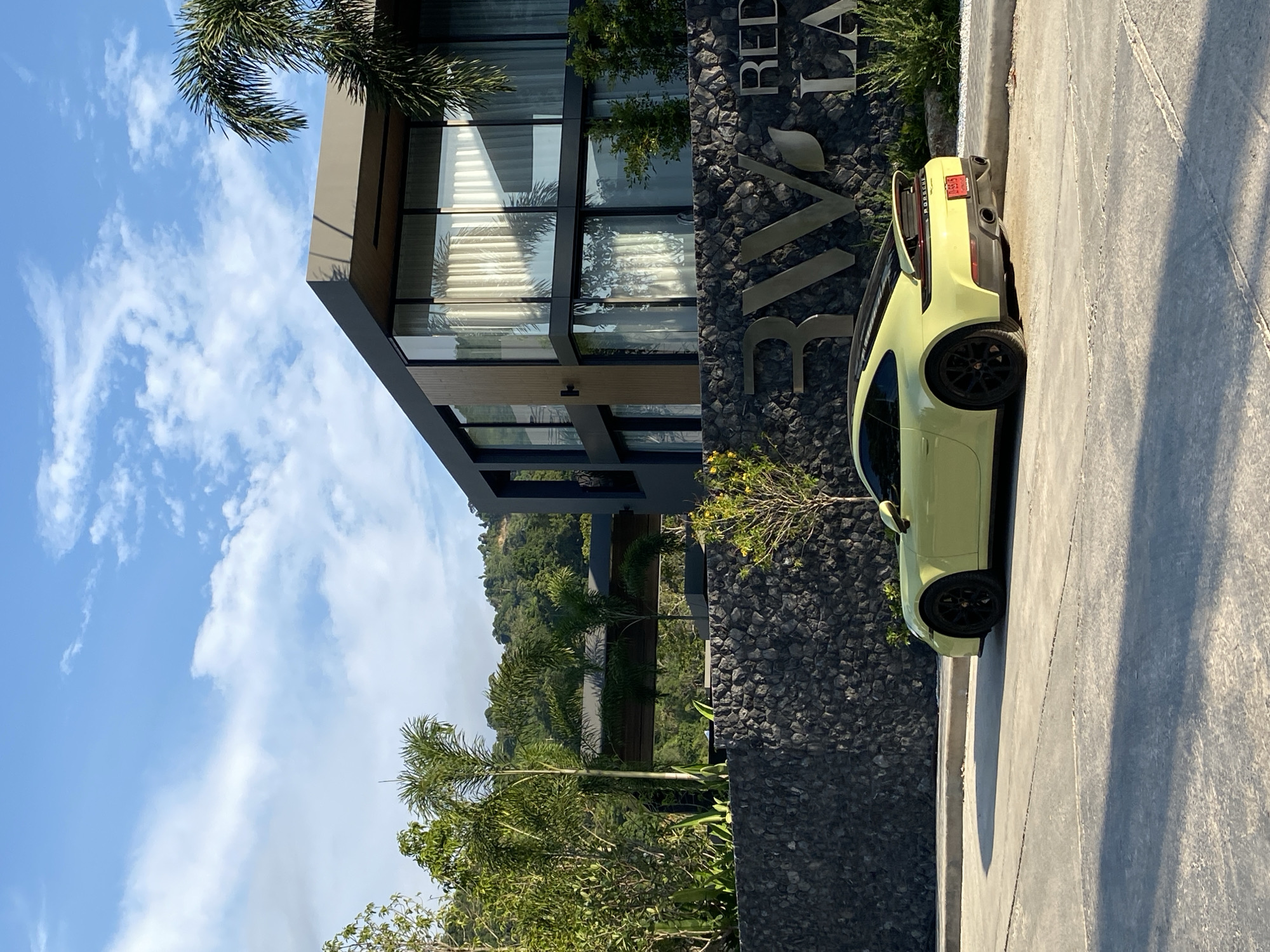 The image depicts a light green Porsche parked next to a building with a dark gray facade adorned with large, metallic lettering that reads "RED LAVA," suggesting this is the name of the building. The building has modern architectural elements, including large glass windows reflecting the sky and surrounding foliage.
The location is suggested to be Choeng Thale, Thailand, based on the prompt. The environment includes lush greenery, palm trees, and a bright blue sky with scattered clouds, indicative of a tropical climate. The car is parked on a light gray pavement or driveway that appears to be newly constructed. The overall scene conveys a sense of luxury and contemporary design, likely representing a high-end residence or resort. No people are visible in the image.
