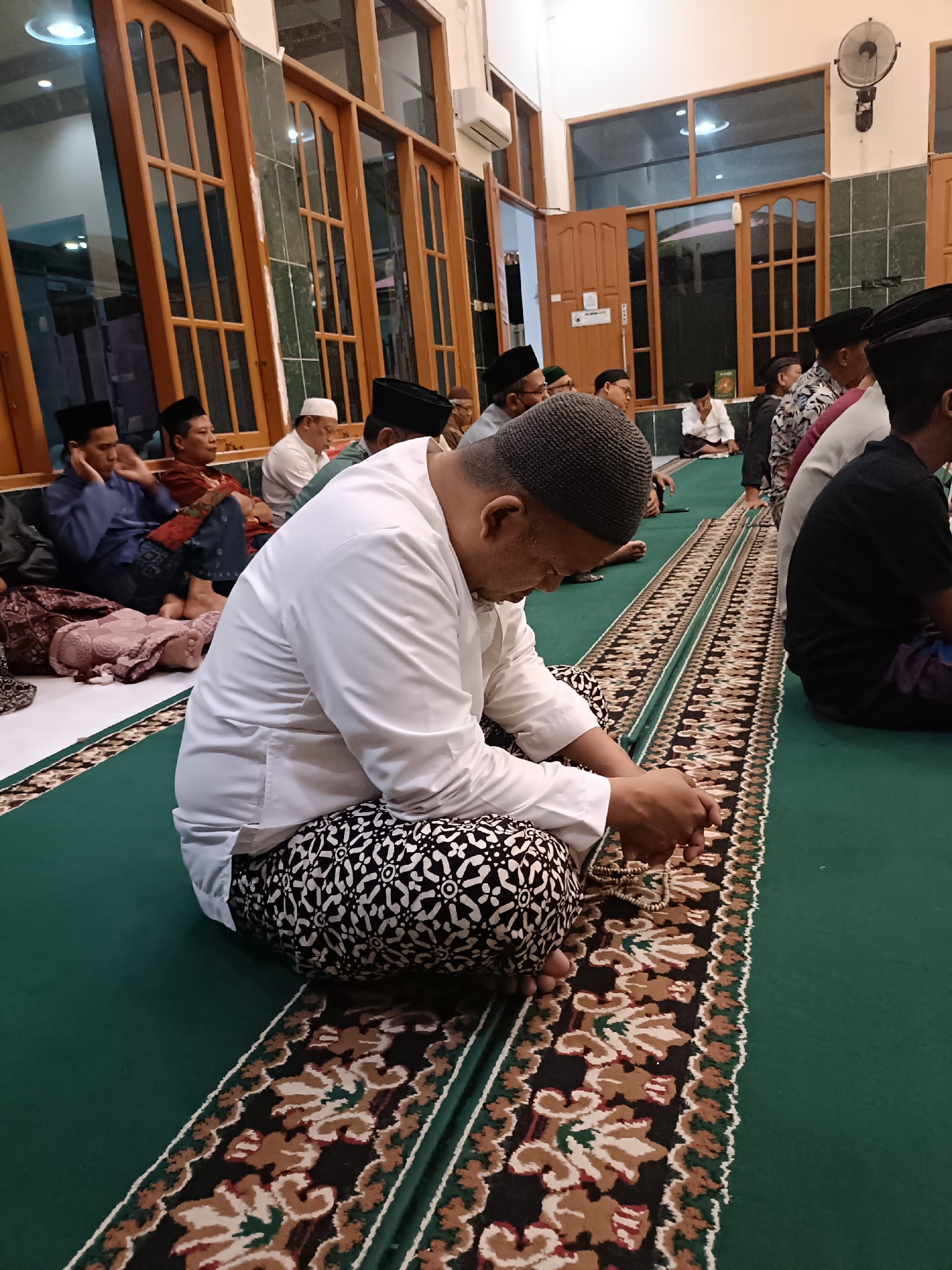 The image depicts an indoor scene, likely a mosque, with several men seated on green carpeted floors. In the foreground, a man wearing a white long-sleeved shirt, a dark skullcap, and patterned pants is seated cross-legged, holding a string of prayer beads. He appears to be engaged in prayer or reflection. Behind him, other men are also seated, some facing forward, others in profile. The room has large wooden-framed windows with glass panes, and a patterned carpet runs along the length of the space. The lighting suggests it is either evening or an overcast day. The presence of prayer beads and the attire of the men strongly indicate a religious gathering, common in mosques in cities like Kediri, Indonesia.