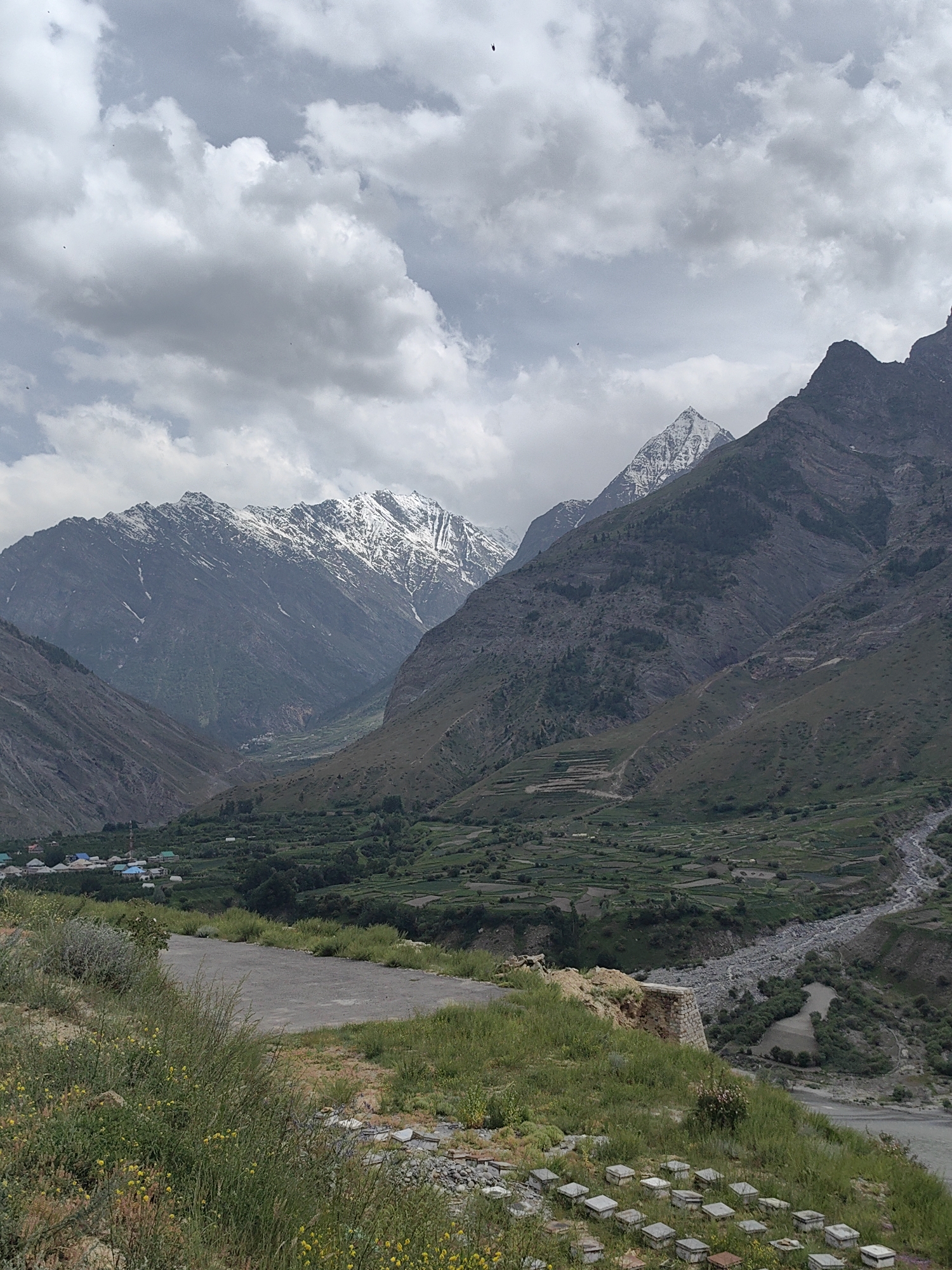 The image depicts a mountainous landscape in Tandi, India. In the foreground, a dirt road is visible, bordered by grassy slopes dotted with small yellow flowers and some scattered grey boxes. The middle ground reveals a valley with terraced fields, a village with houses featuring colorful roofs, and a winding riverbed. The background is dominated by imposing, dark mountains, some of which are capped with snow. The sky is overcast with a mix of grey and white clouds. Several birds are visible as small dark specks in the sky.