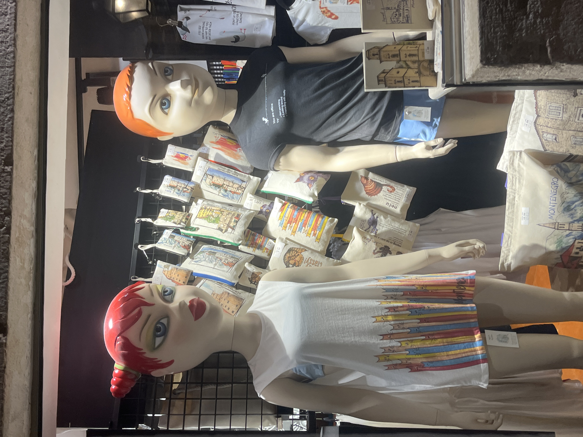 In Budva, Montenegro, a retail display features two mannequins. The mannequin on the left, with red hair styled in pigtails and prominent makeup, wears a white sleeveless dress adorned with a colorful pattern of standing cats and the word "Montenegro". The mannequin on the right, with orange hair, wears a black t-shirt with white text that reads "You are offline" and then lists several possible actions, followed by "Looking out of the window" and "Interacting with reality". This mannequin also wears blue shorts. Behind the mannequins, a rack displays numerous small bags or pouches with various illustrated designs, including cats and architectural scenes. To the right of the right mannequin, a framed artwork depicting a building is visible. In the foreground, several tote bags with similar illustrated designs, including one with the word "Montenegro" printed on it, are displayed. The background is dark, with some shelving and merchandise visible. The overall scene suggests a souvenir or clothing shop.