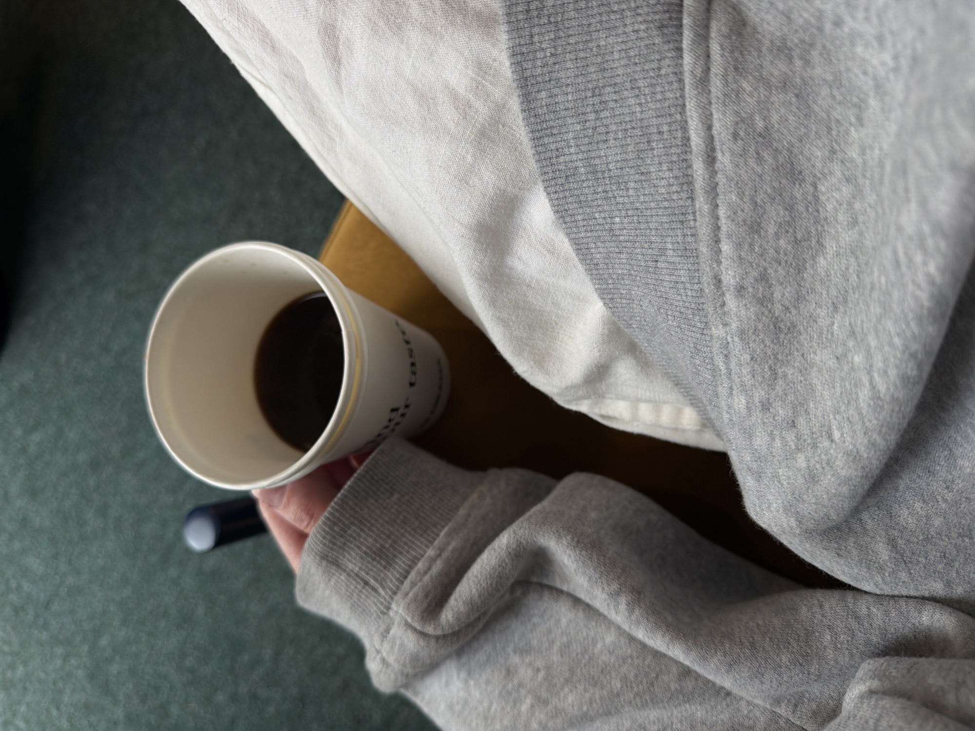 A close-up, top-down shot captures a person's hand, clad in a grey long-sleeved sweatshirt or hoodie with a ribbed cuff, holding a white paper coffee cup. The cup contains a dark liquid, likely coffee, and features partial text on its side that appears to read "tast" or "past."
The scene is an indoor setting. The hand and cup are positioned over a light brown surface, possibly a table or the person's lap. To the upper right, a section of lighter, off-white fabric with a distinct linen-like weave is visible, draped over the grey garment. The lower left portion of the image reveals a dark green, textured surface, likely a carpeted floor. A dark cylindrical object, resembling a pen or marker with a light tip, lies on this surface near the person's hand.
The primary activity is holding and presumably drinking or about to drink coffee. The overall impression is one of relaxed comfort, perhaps during a casual break in Tbilisi, Georgia. The lighting is soft and ambient, characteristic of an indoor environment. No specific time of day, weather, or emotions are discernible from the image.