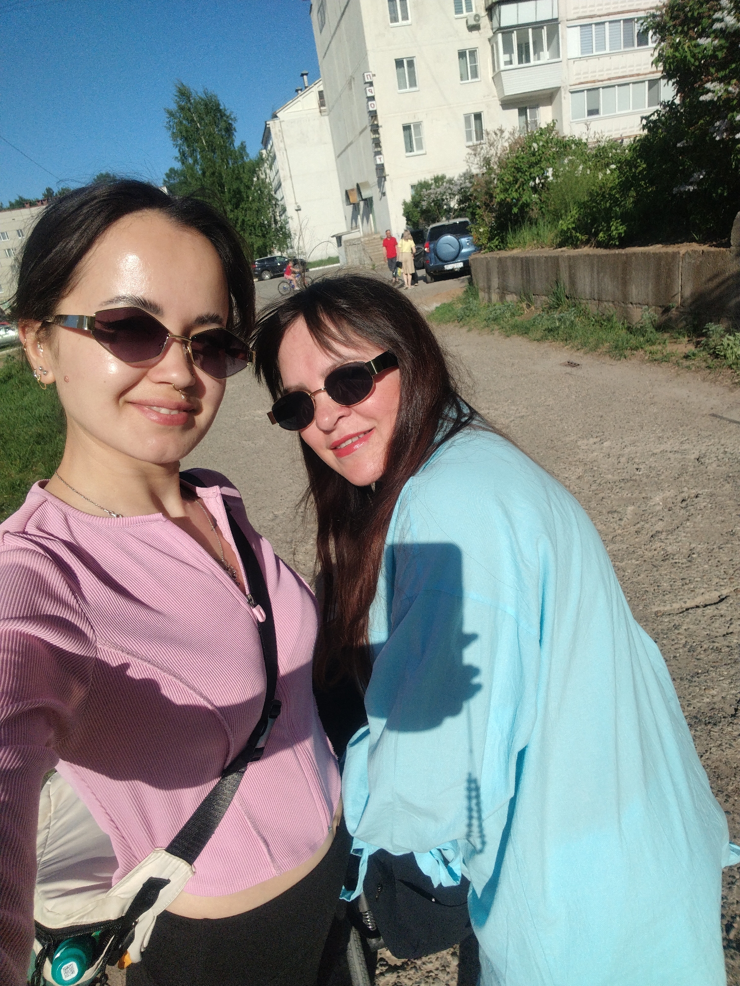 Two women pose for a selfie on a paved walkway. The woman on the left wears a pink ribbed top, sunglasses, and a crossbody bag. The woman on the right wears a light blue blouse and sunglasses. In the background, a multistory building stands with visible windows. A blue SUV is parked near the building, and two individuals stand beside it. A bicycle is visible in the background. The location is Noviy, Russia.