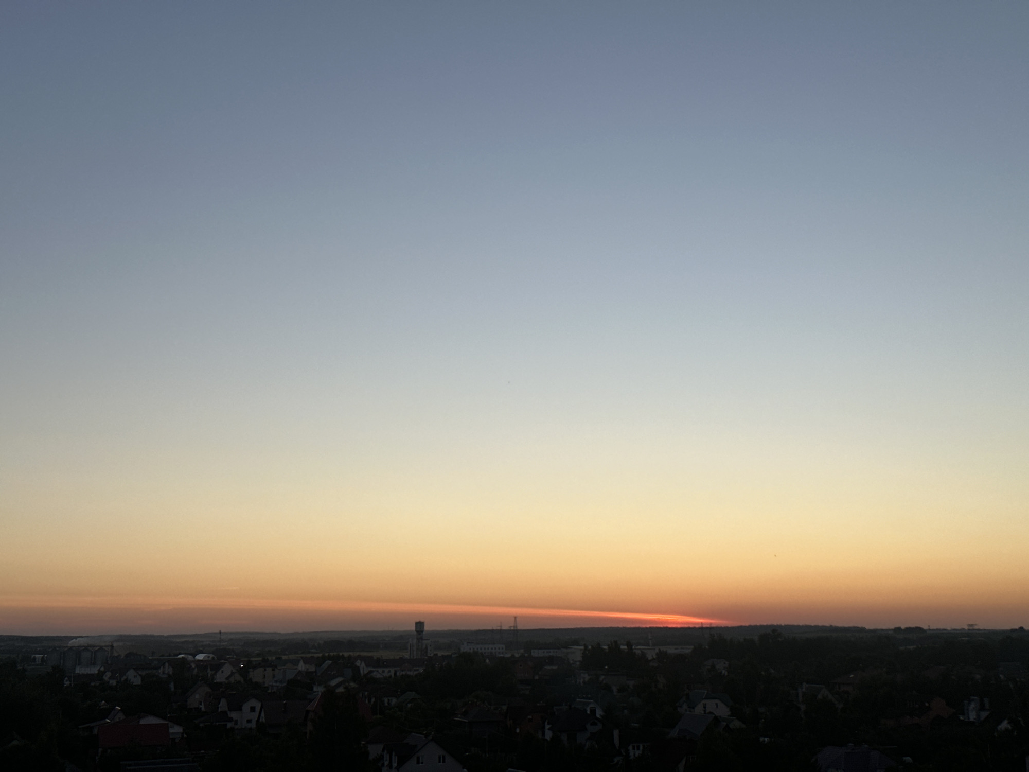 The image captures a panoramic view of a suburban or rural landscape at dusk, likely in the vicinity of Шчомысліцкі сельскі Савет, Belarus. The sky displays a gradient of colors, transitioning from a pale blue at the top to a warm orange and red hue near the horizon, indicating sunset. Below the sky, a silhouetted town or village is visible, characterized by a collection of rooftops and scattered trees. A tall, slender structure, possibly a water tower or radio mast, stands out against the darkening landscape in the middle distance. The overall impression is one of tranquility and the end of a day.