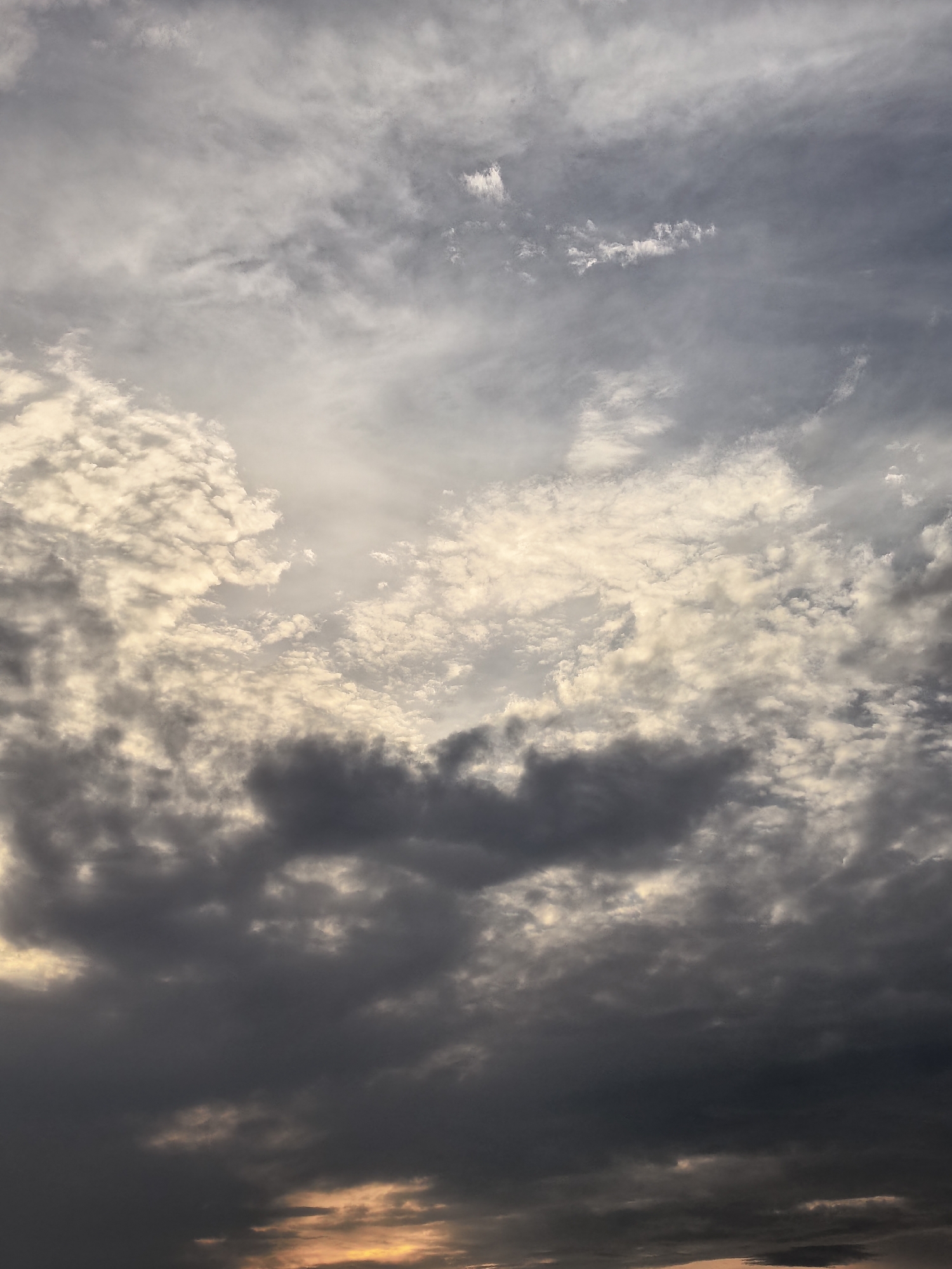This is an image of a sky filled with a variety of clouds, appearing to be taken during sunset or sunrise. The clouds range in color from dark grey and ominous to light and wispy, with patches of golden light peeking through in the lower portion of the image. The overall impression is one of dramatic and dynamic weather. The location is unknown.