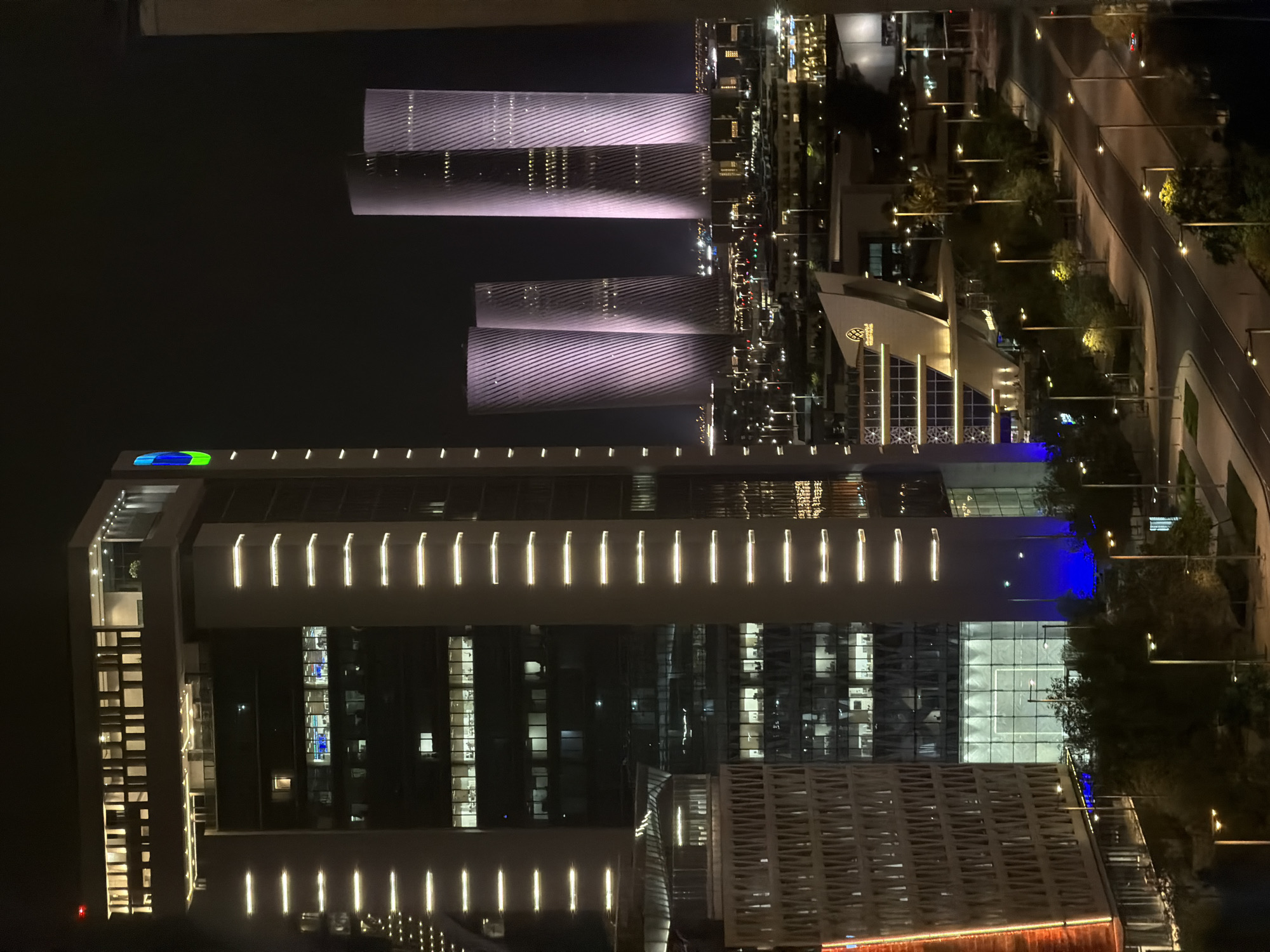 The image depicts a nighttime cityscape with a focus on several modern skyscrapers. In the foreground, a tall, rectangular building is illuminated with vertical lines of white lights on its facade. This building has multiple floors with visible interior lighting, suggesting office spaces. To its left, another building with a more complex, textured facade and orange lighting is partially visible.
Further in the background, a cluster of three distinct cylindrical towers stands out. These towers are lit with a purple hue and feature a patterned texture on their surfaces, possibly indicating architectural design elements or lighting effects. The cityscape extends into the distance with more buildings and lights, suggesting a densely populated urban area. A road with streetlights and sparse vehicle traffic is visible in the lower portion of the image. Trees line the road, and some of the background buildings have a more traditional architectural style, contrasting with the modern towers. The sky is dark, characteristic of nighttime.