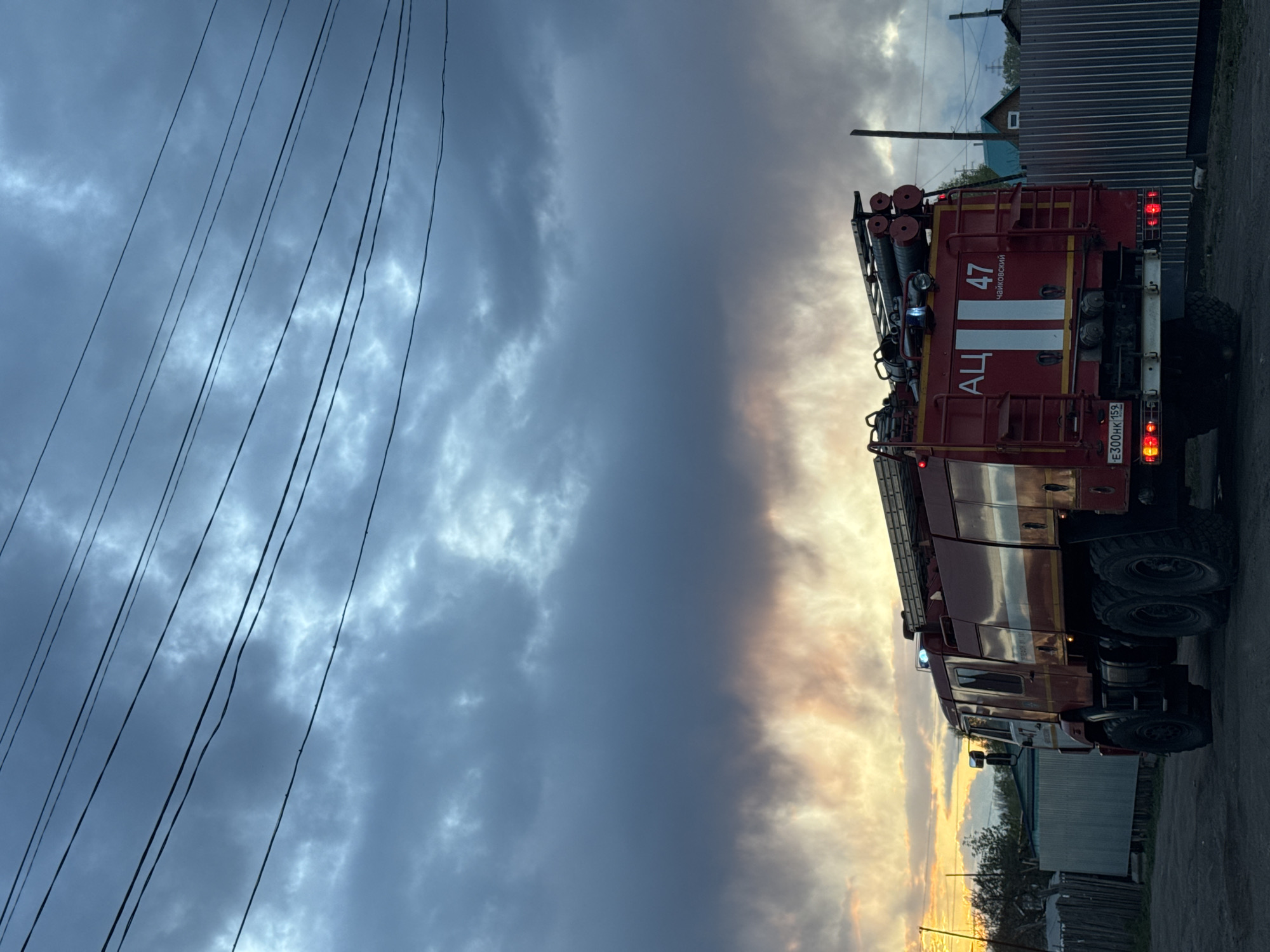 The image shows a fire truck parked on a street in what appears to be a residential area of Chaikovsky, Russia. The fire truck is red and has the number "47" and the Cyrillic letters "АЦ" (ATs) on its side, which likely stands for "Автоцистерна" (Avtotsisterna), meaning "tanker truck". The license plate is visible and reads "E300HK 159". The sky above is cloudy and ominous, with a mixture of dark and golden-tinged clouds, possibly indicating a recent sunset. Utility lines are visible overhead. The environment consists of simple buildings, possibly houses or sheds, and a fence, suggestive of a typical small-town or rural setting. There are no people visible in the scene.