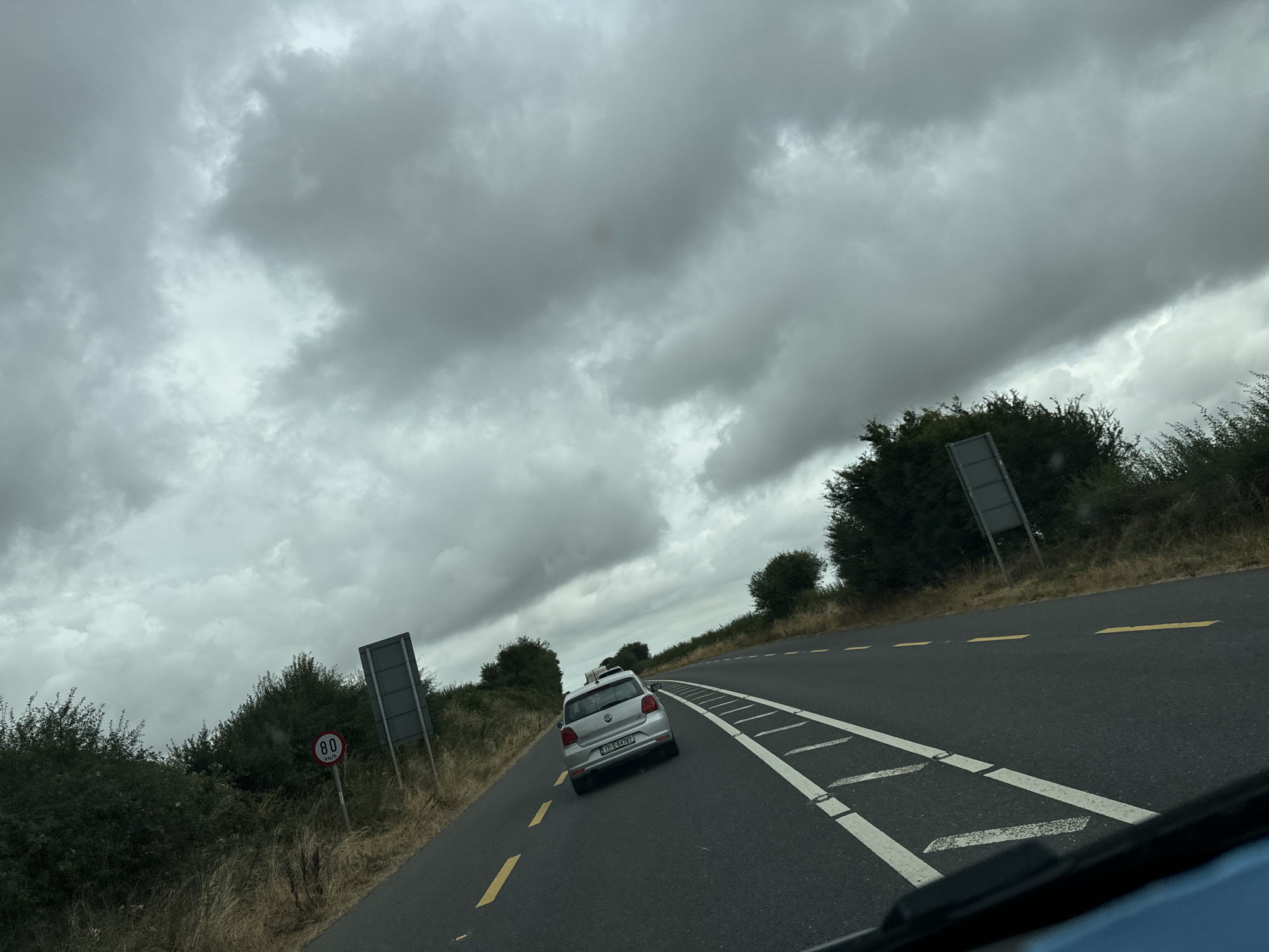 The image shows a view from inside a vehicle traveling on a two-lane road in Ireland. The sky is heavily overcast with dark, dramatic clouds. On the left side of the road, there is a speed limit sign indicating 80 km/h, and an empty, tall grey signpost. A dense line of green trees and bushes borders the road on both sides. A silver Volkswagen hatchback with a luggage rack on top is in front of the vehicle, in the right lane. The road is asphalt with yellow lines marking the lanes and white dashed lines indicating the edge of the travel lane. The overall impression is of a rural or semi-rural highway under a cloudy sky.