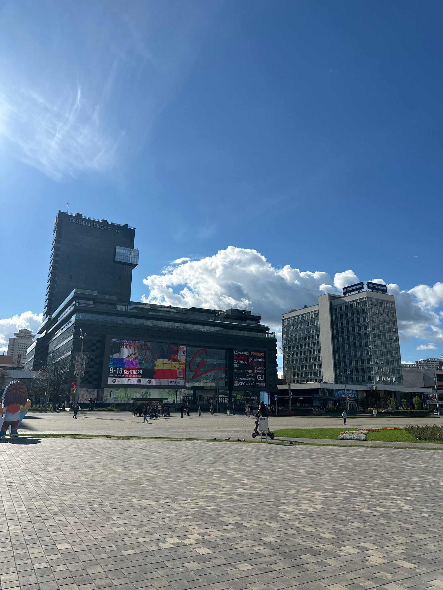 The image shows a wide-angle view of a city square in Minsk, Belarus, on a sunny day with scattered clouds. In the foreground, a paved plaza with a geometric pattern of grey and darker grey square tiles is visible. Several people are walking or standing in the mid-ground, and a person is riding an electric scooter. To the left, a large, colorful, cartoonish statue of a creature with shaggy brown hair and a blue shirt is present.
Dominating the background are several buildings. The most prominent is a dark, modern, high-rise building with "DoubleTree by Hilton" signage at the top. Below this building is a multi-story structure with a large digital billboard displaying advertisements and event information, including text in Cyrillic that reads "9-13 СЕНТЯБРЯ" (September 9-13) and "МИНСК" (Minsk). A lighter-colored, older, multi-story office building stands to the right of the DoubleTree hotel, with "БЕЛТЕЛЕКОМ" (BELTELECOM) signage on its roof. The area in front of these buildings features a green lawn with flowerbeds and some trees. The sky is bright blue with wispy white clouds and the sun is shining from the left side of the frame, casting shadows. The scene depicts an urban public space with commercial and hospitality establishments.