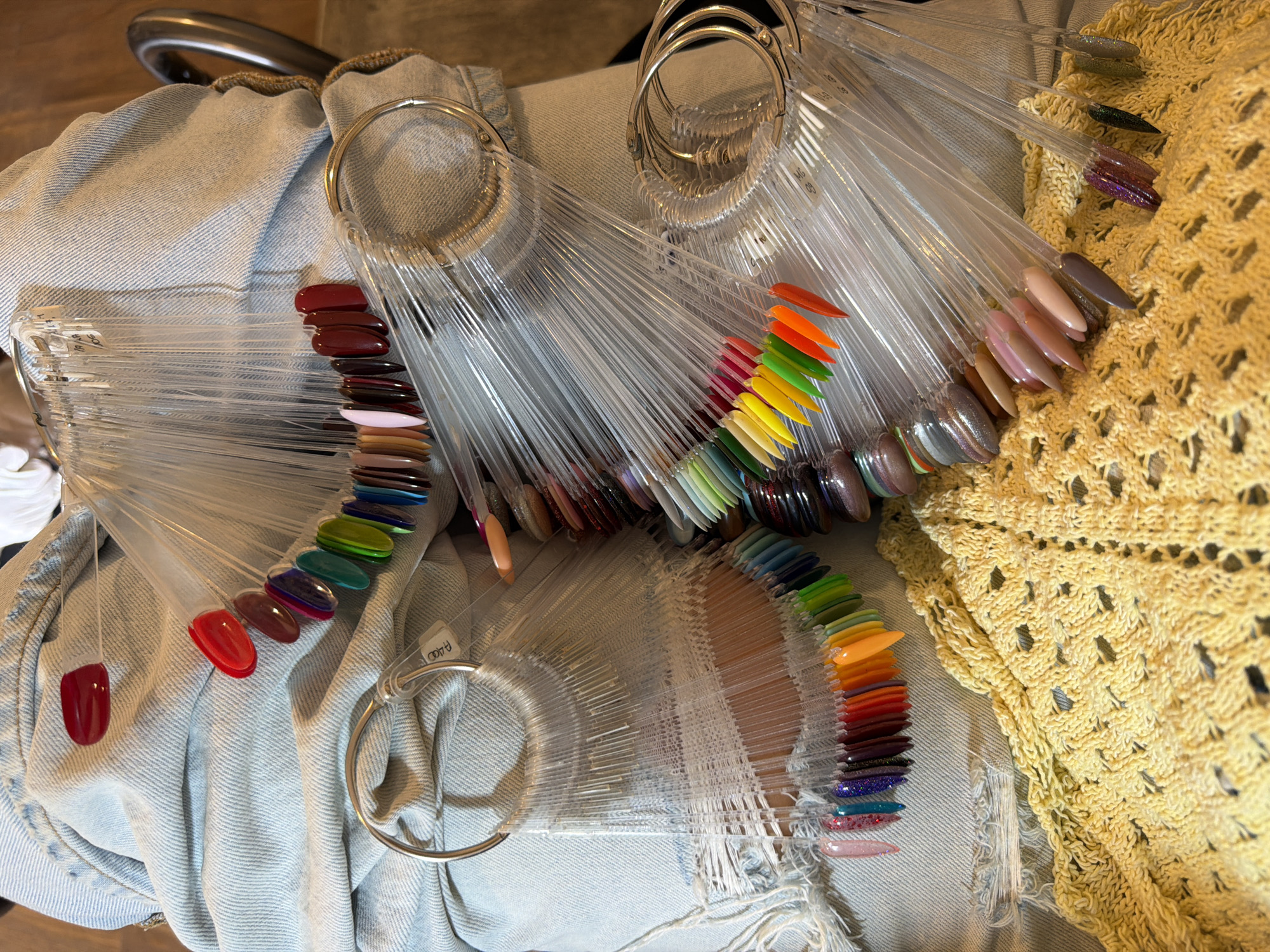 In North Kuta, Indonesia, a person is seated on a chair, wearing light blue denim jeans that are ripped at the knee. Their lap is covered with various color swatch fans for nail polish. There are at least three distinct fans visible, each holding numerous individual nail polish samples on plastic tips. The samples range in color from bright reds, oranges, yellows, and greens to more muted browns, pinks, and purples. Some of the nail polish samples appear to have glitter or a metallic finish. A yellow knitted garment is draped over the lower portion of the person's lap. The background is blurred but suggests an indoor setting.