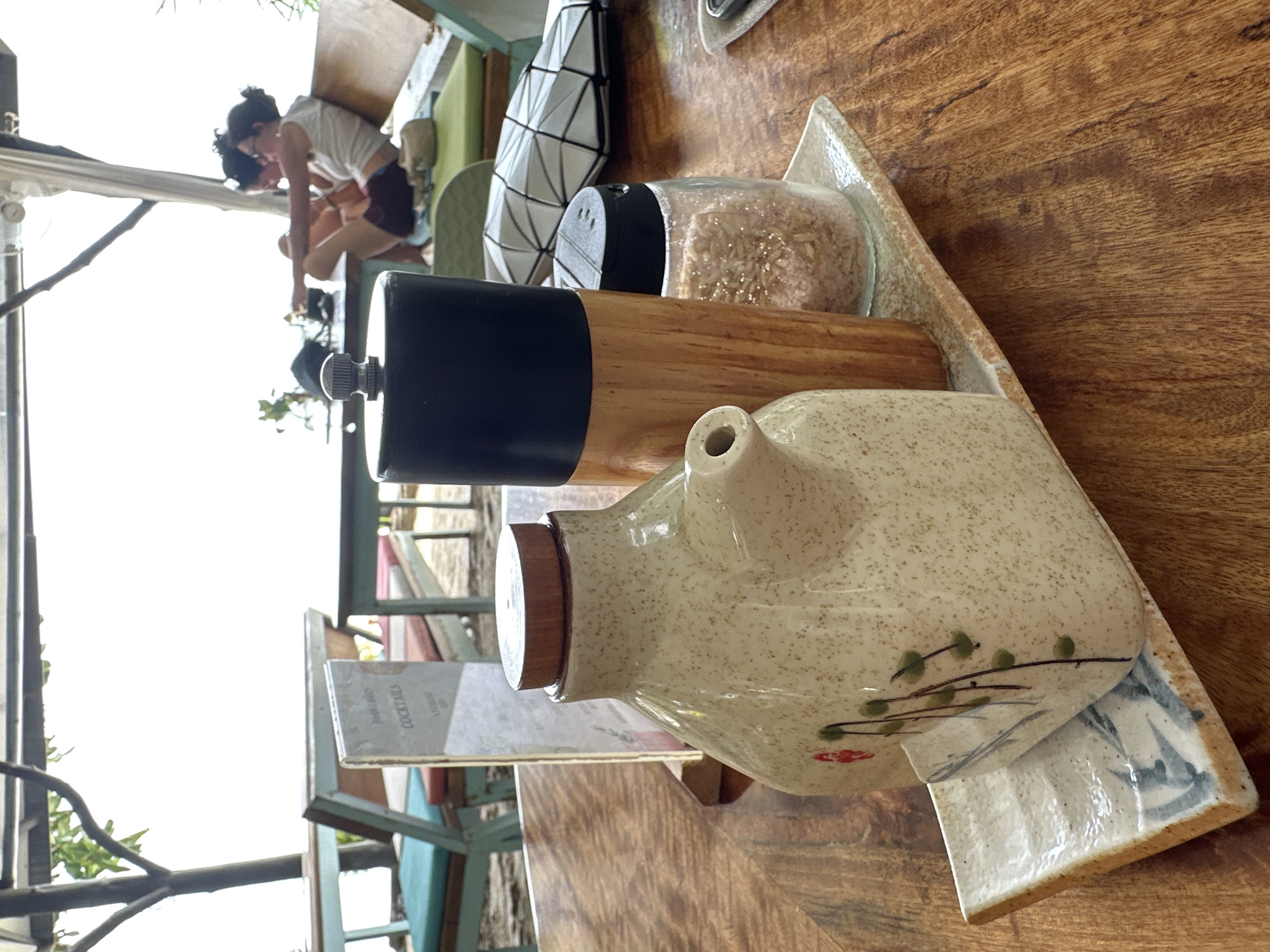 In Ko Pha-ngan District, Thailand, on a wooden table outdoors, a ceramic dispenser with a wooden lid and a floral design sits on a decorative rectangular plate. Next to it are a wooden and metal pepper grinder and a glass jar filled with coarse pink salt. In the background, a couple sits on a wooden bench in a shaded area, with a white, geometric patterned bag placed on the bench. Further back, there are tables and chairs set up on a sandy surface. The scene appears to be at a beachside restaurant or cafe.