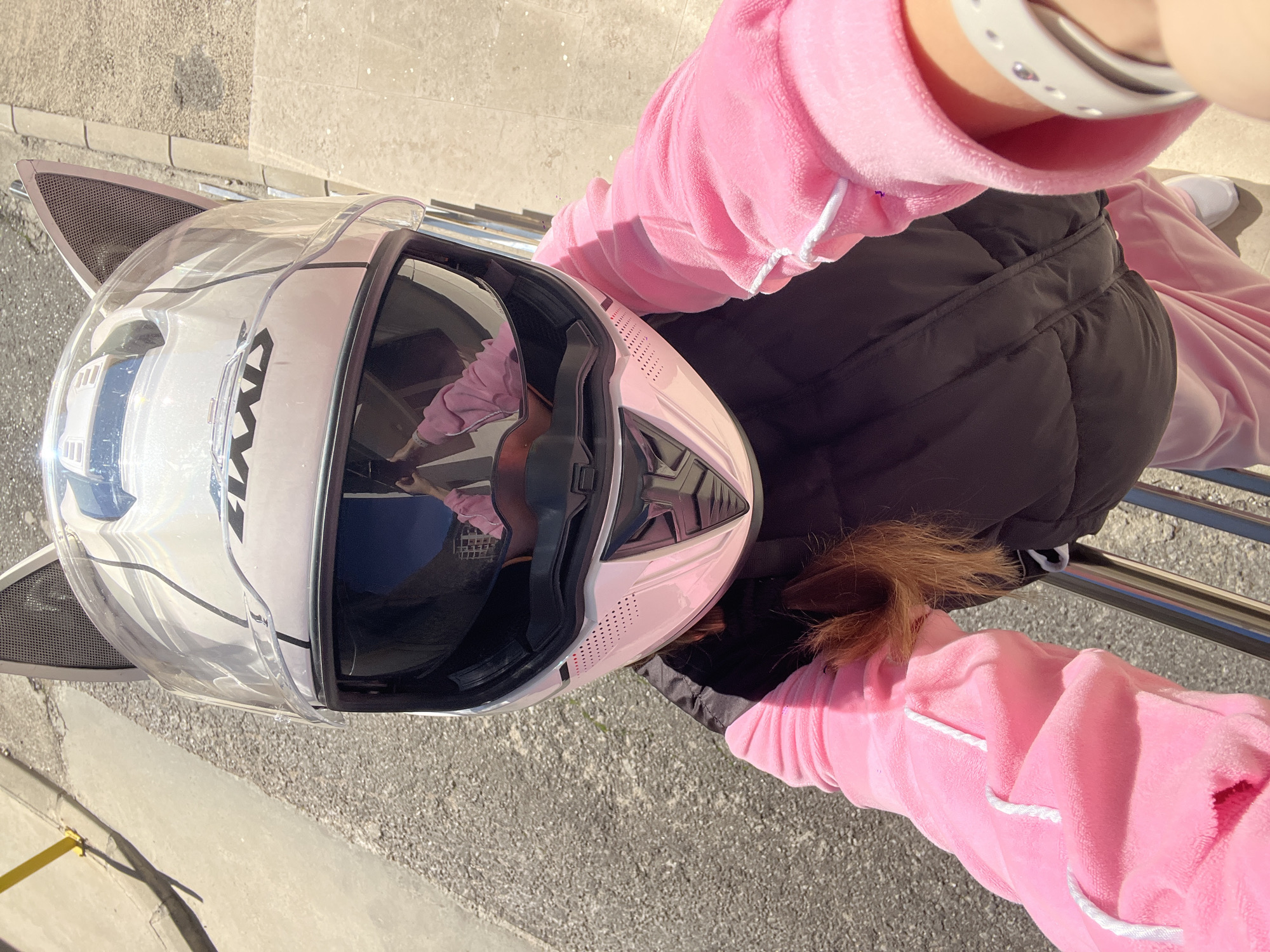 A top-down, medium shot depicts a person seated outdoors, likely in Rafailovići, Montenegro, though specific location details are not directly visible in the image. The individual wears a light pink velvet sweatsuit with white trim and a black puffer vest. The person is also wearing a white smartwatch. A white motorcycle helmet with attached cat ears sits beside the person on a metal railing. The surface below the person is concrete.