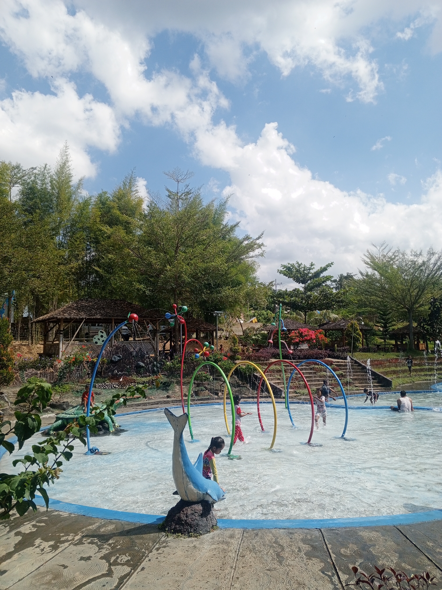 The image depicts a lively water play area in Darsono, Indonesia, under a bright, partly cloudy sky. In the foreground, a shallow pool is filled with clear water, featuring colorful archways that spray water. Children are actively engaged in the water, some running and splashing, while others sit or stand near the spray arches. A blue dolphin statue on a rock is positioned prominently to the left. Beyond the water area, a tiered landscape rises, with various plants and trees, including bamboo, creating a lush backdrop. Traditional Indonesian-style huts with thatched roofs are nestled into the hillside, providing shade. In the distance, more greenery and trees are visible, suggesting a natural, park-like setting. The overall atmosphere is one of recreation and enjoyment on a sunny day.