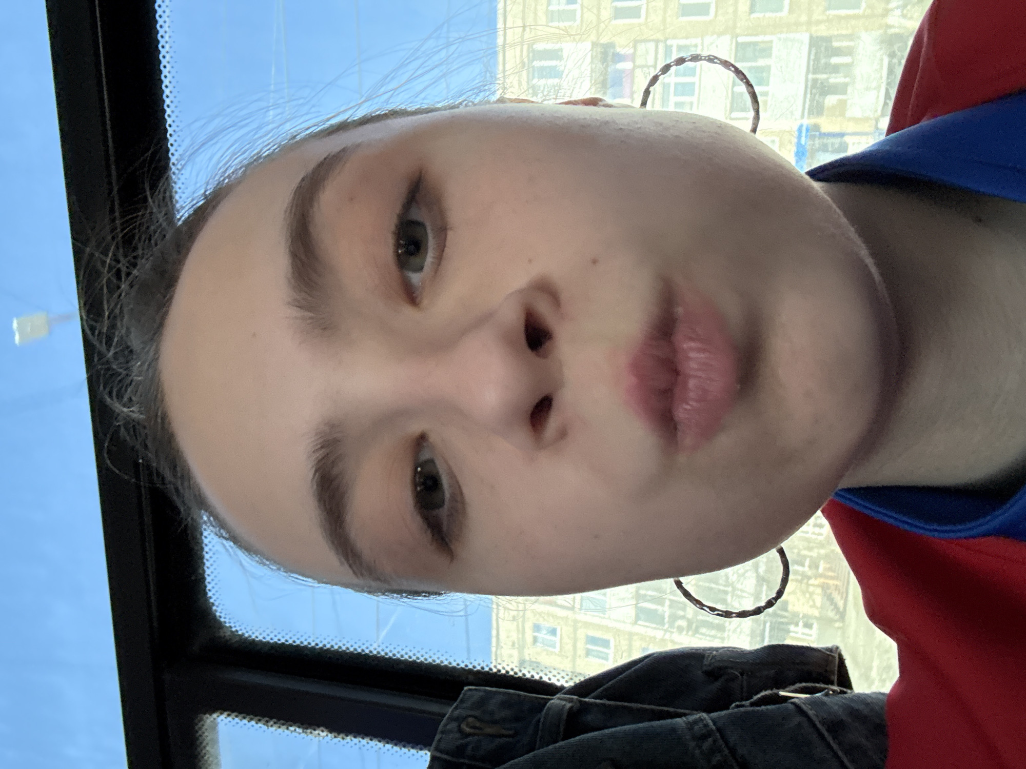 A close-up, upward-tilted selfie captures a person with light skin, full lips pursed slightly, and light-colored eyes, likely inside a vehicle. The window frame and a glimpse of blue sky are visible on the left. Behind the person, through the window, a multi-story building with multiple windows is discernible. The individual wears hoop earrings and a garment with red and blue fabric. A dark denim jacket rests partially in the lower portion of the frame. The location is unknown.