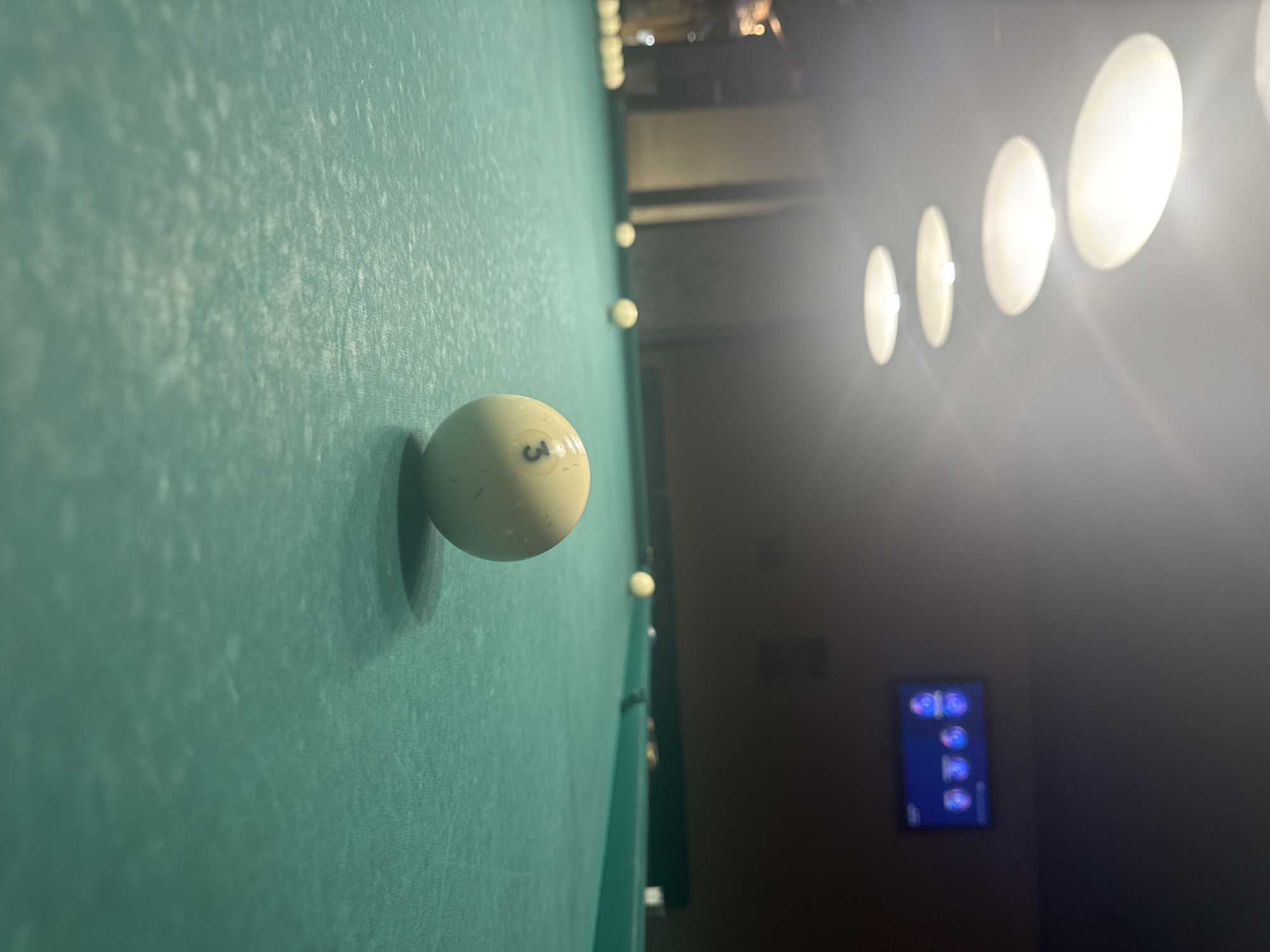 The scene depicts a billiard table with a green felt surface. A yellow billiard ball, marked with the number "3," rests on the table's surface in the foreground. Multiple other balls are visible along the far side of the table. Above the table, a series of round lights are illuminating the scene. In the background, a television screen displays an image with blue and purple hues. The location in the city of Moscow, Russia is not apparent from the image.