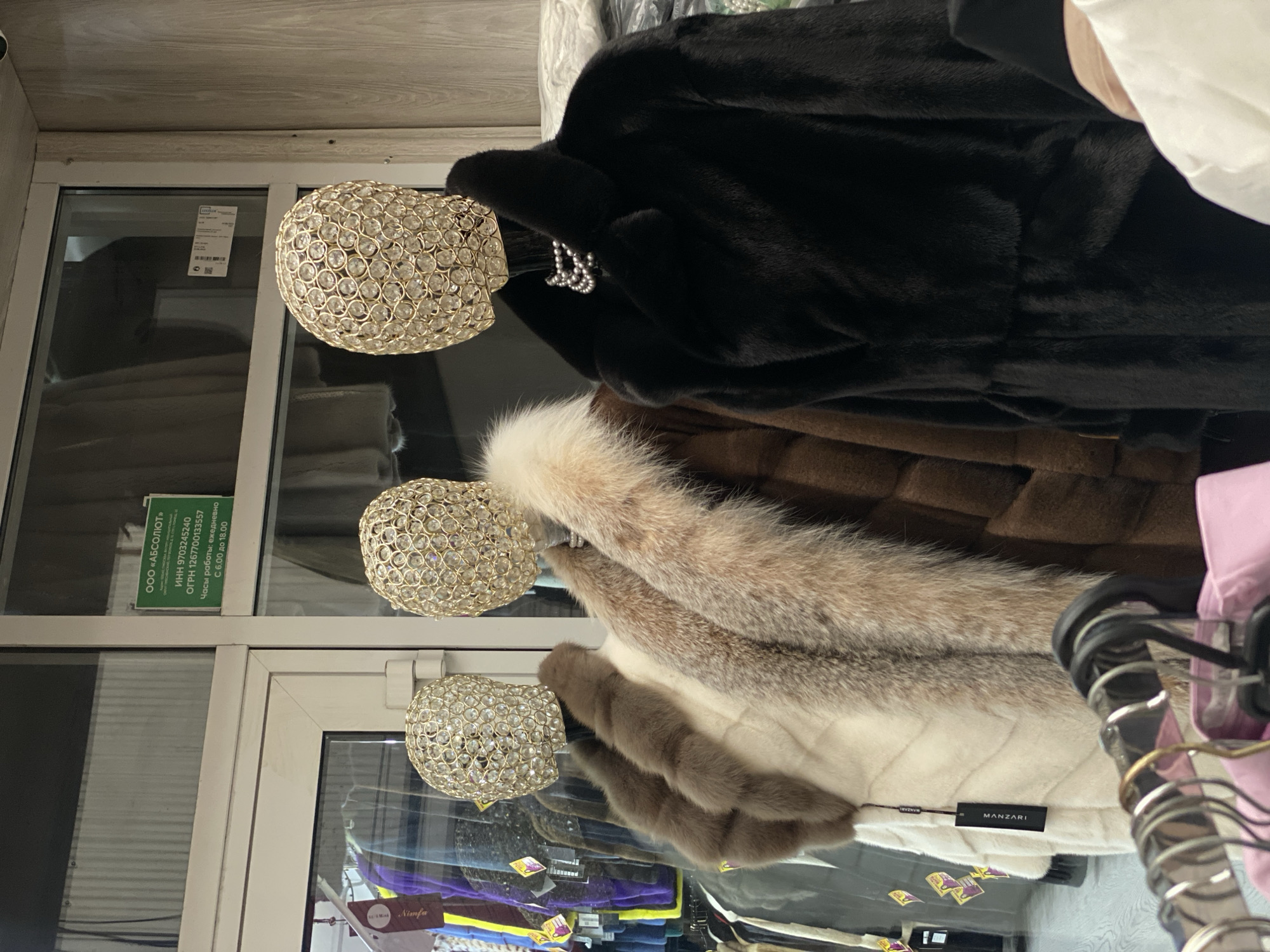 In Moscow, Russia, at a retail establishment, three mannequins are displayed in the foreground. The mannequins are adorned with luxurious fur coats and have distinctive, ornate, crystal-encrusted heads. The leftmost mannequin wears a white fur coat with brown fur trim. The middle mannequin is dressed in a brown fur coat. The rightmost mannequin is presented in a long, black fur coat, accessorized with a pearl necklace. Behind the mannequins, various other garments are visible, including what appear to be colorful scarves or accessories on the left, and more fur coats on racks. A green sign with Russian text is affixed to the glass partition on the upper left of the image, indicating store hours and company information. The overall environment suggests a high-end clothing store specializing in fur apparel.