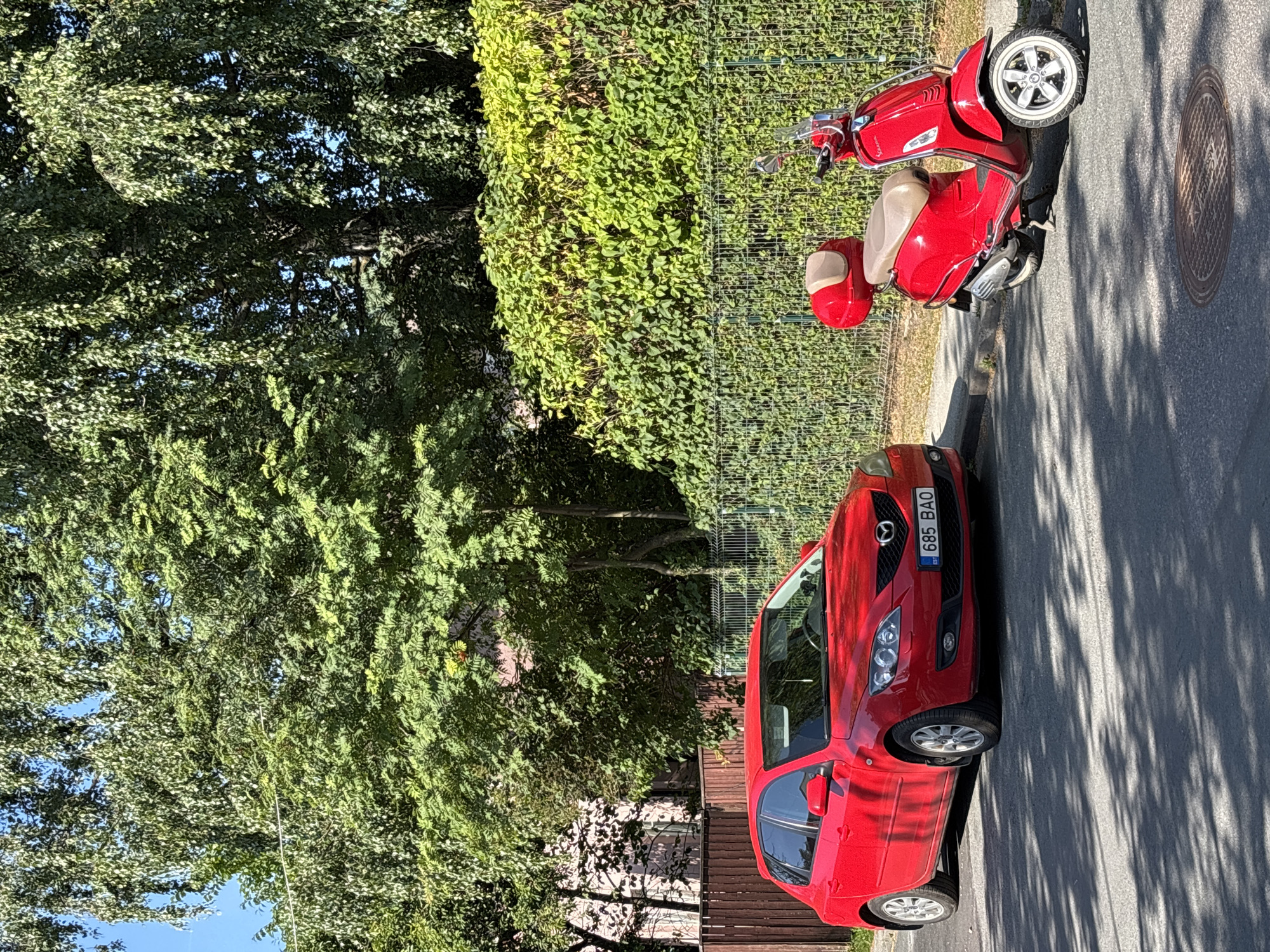 A red hatchback car is parked on the side of a paved street in Tallinn, Estonia. The car's license plate reads "685 BAO". To the right of the car, a red scooter with a beige seat and a red top box is parked. Both vehicles are parked on the right side of the road, facing away from the camera. Behind the vehicles, there is a low wooden fence, followed by a green metal fence, and dense green foliage. Tall trees with lush green leaves are visible in the background, with a few buildings peeking through the trees. The sky is clear and blue. Sunlight casts shadows on the road and the vehicles, indicating a sunny day.